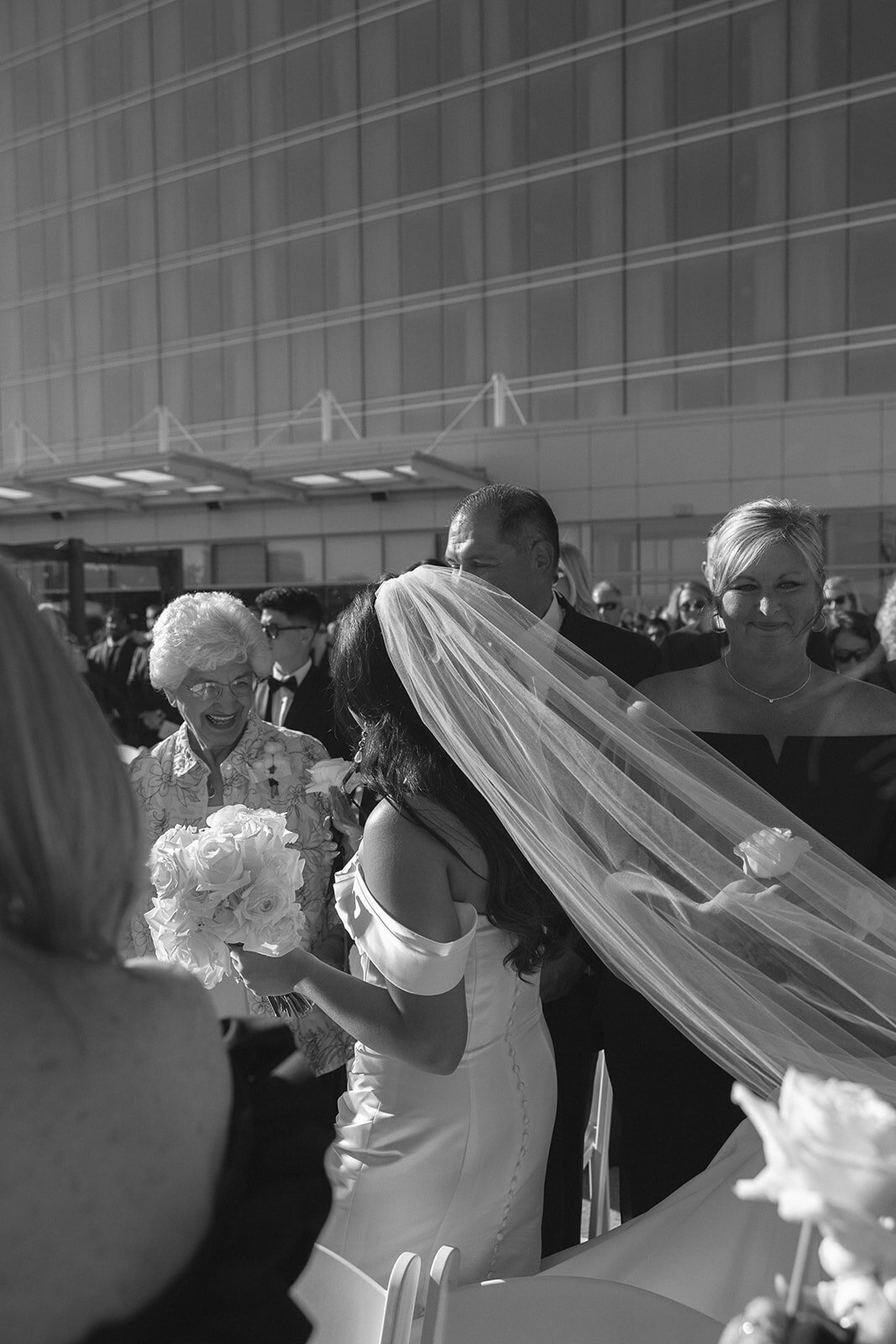 Candid wedding moment in St. Louis — joyful bride in off-shoulder gown and long veil laughing with family during outdoor ceremony, surrounded by loved ones in natural sunlight. Captured by Missouri-based traveling wedding photographer specializing in authentic, emotional, documentary-style wedding photography