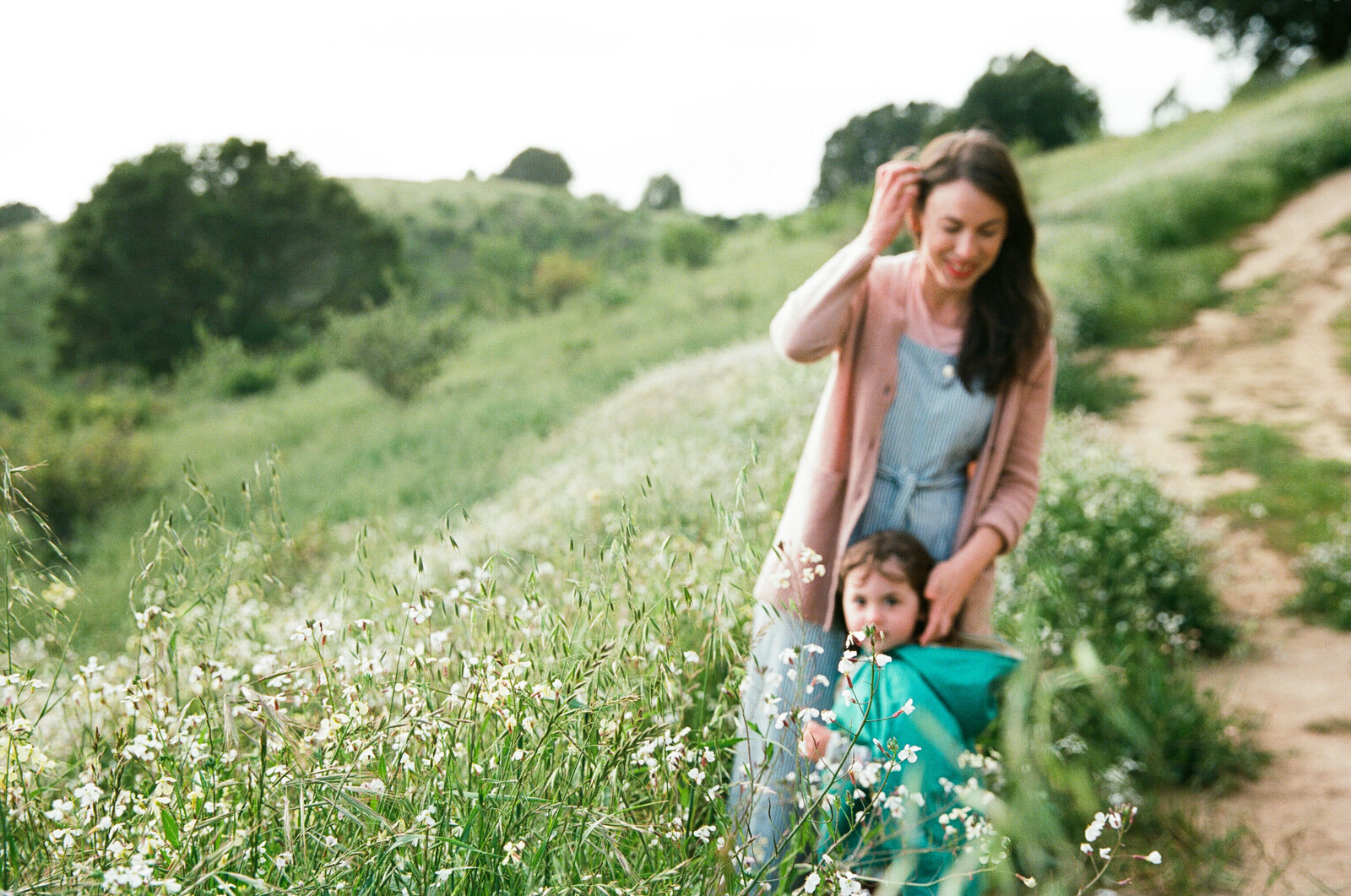 film photograph of a mom in a chambray jumpsuit and blush pink cardigan standing with her young daughter wearing a green cape on a green hillside in oakland california