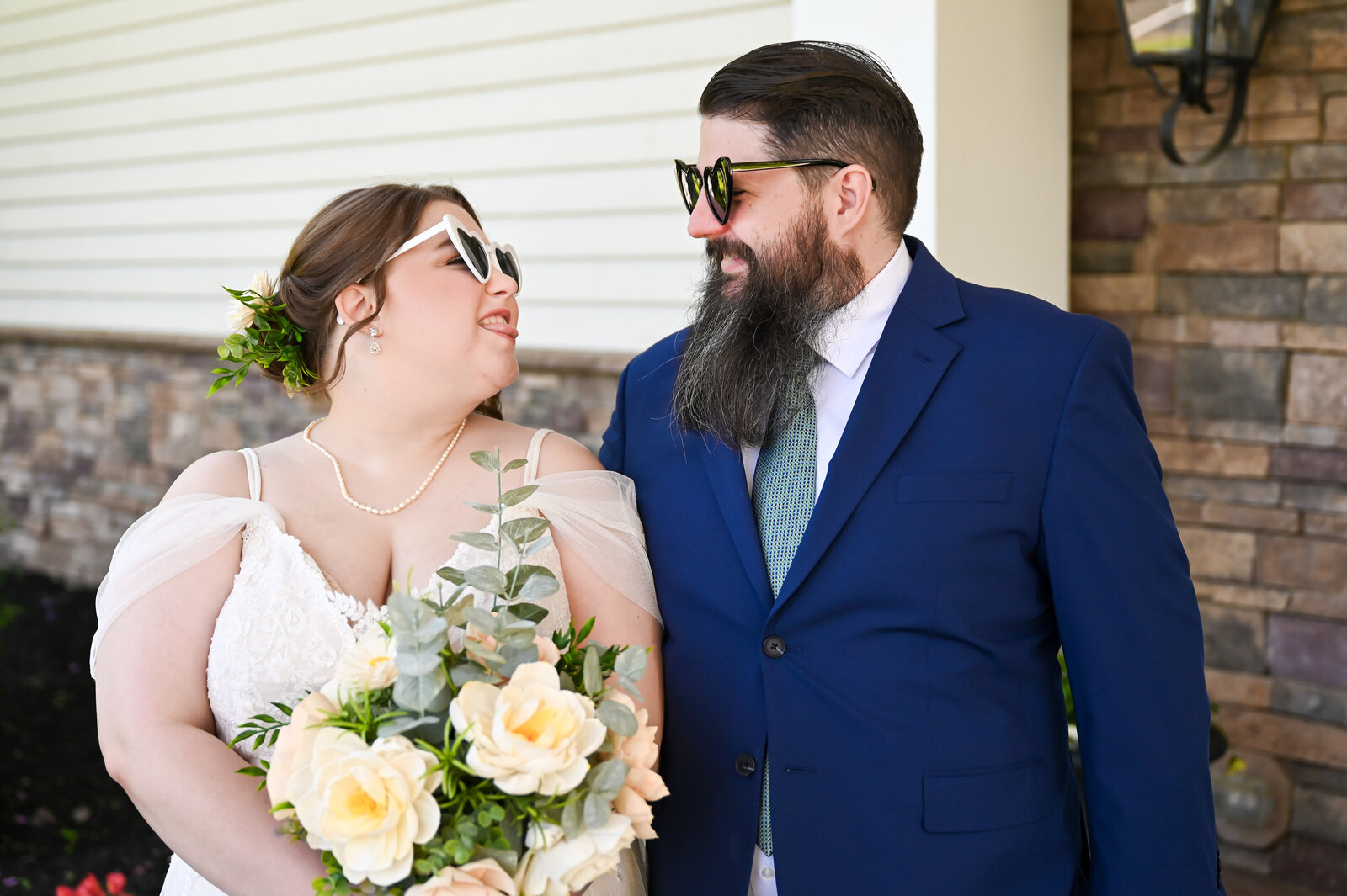 A couple wearing sunglasses smiles at each other. The woman holds a bouquet of yellow flowers. They stand in front of a building with stone and siding.