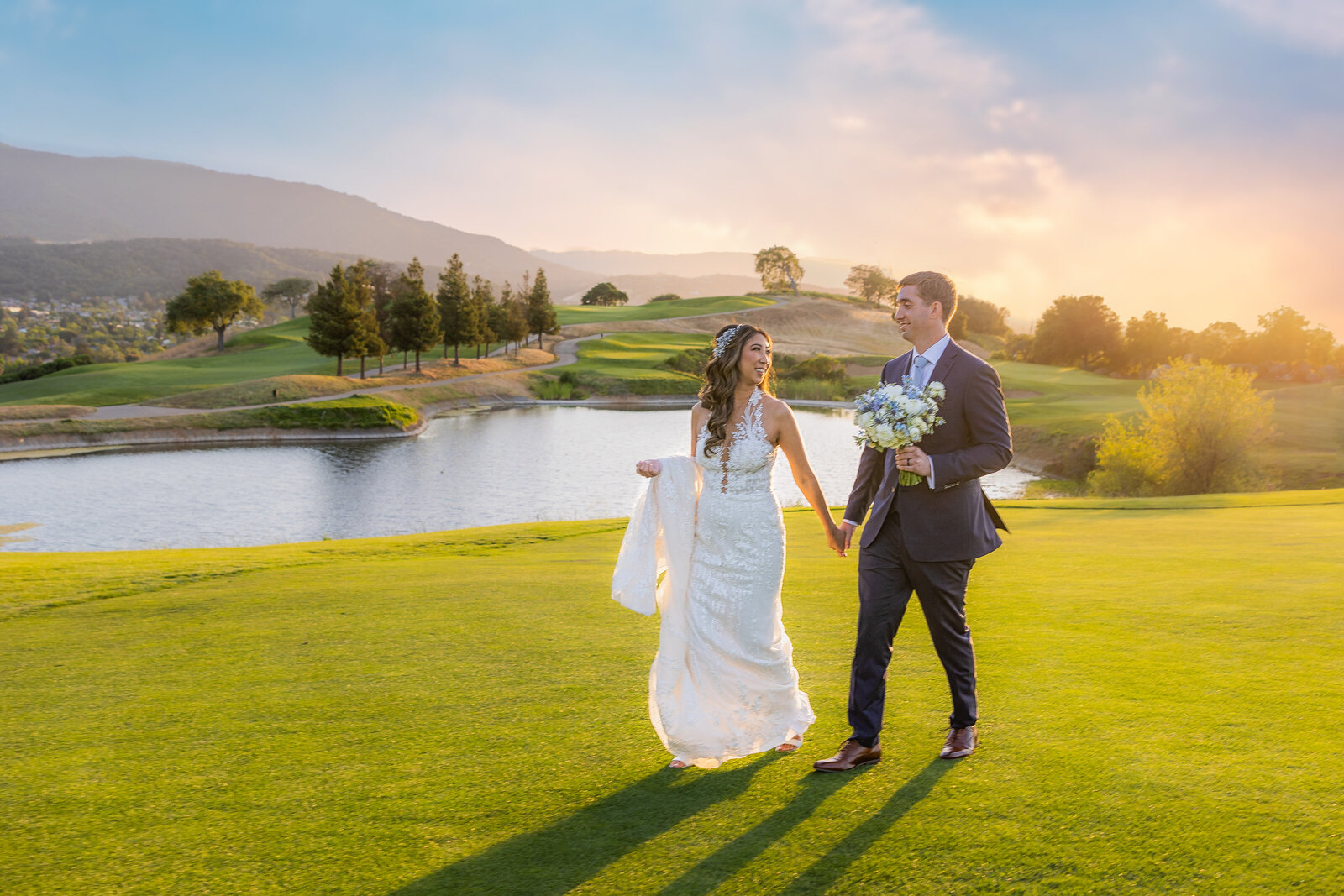 Bride and Groom hold hands as they walk on a golf course with lake in the background and sunset. They look at each other lovingly.  Photo by wedding photographer in Sacramento, Philippe Studio Pro.