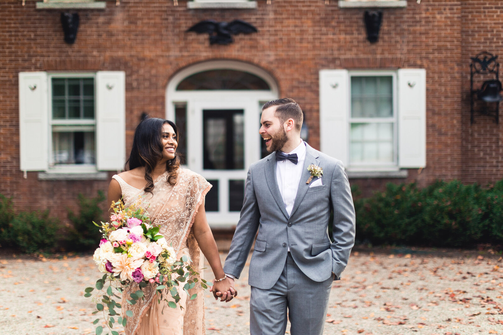 Beautiful Couple in front of brick building. She is in a wedding Sari with a beautiful bouquet and he is in a grey suit and bow tie.