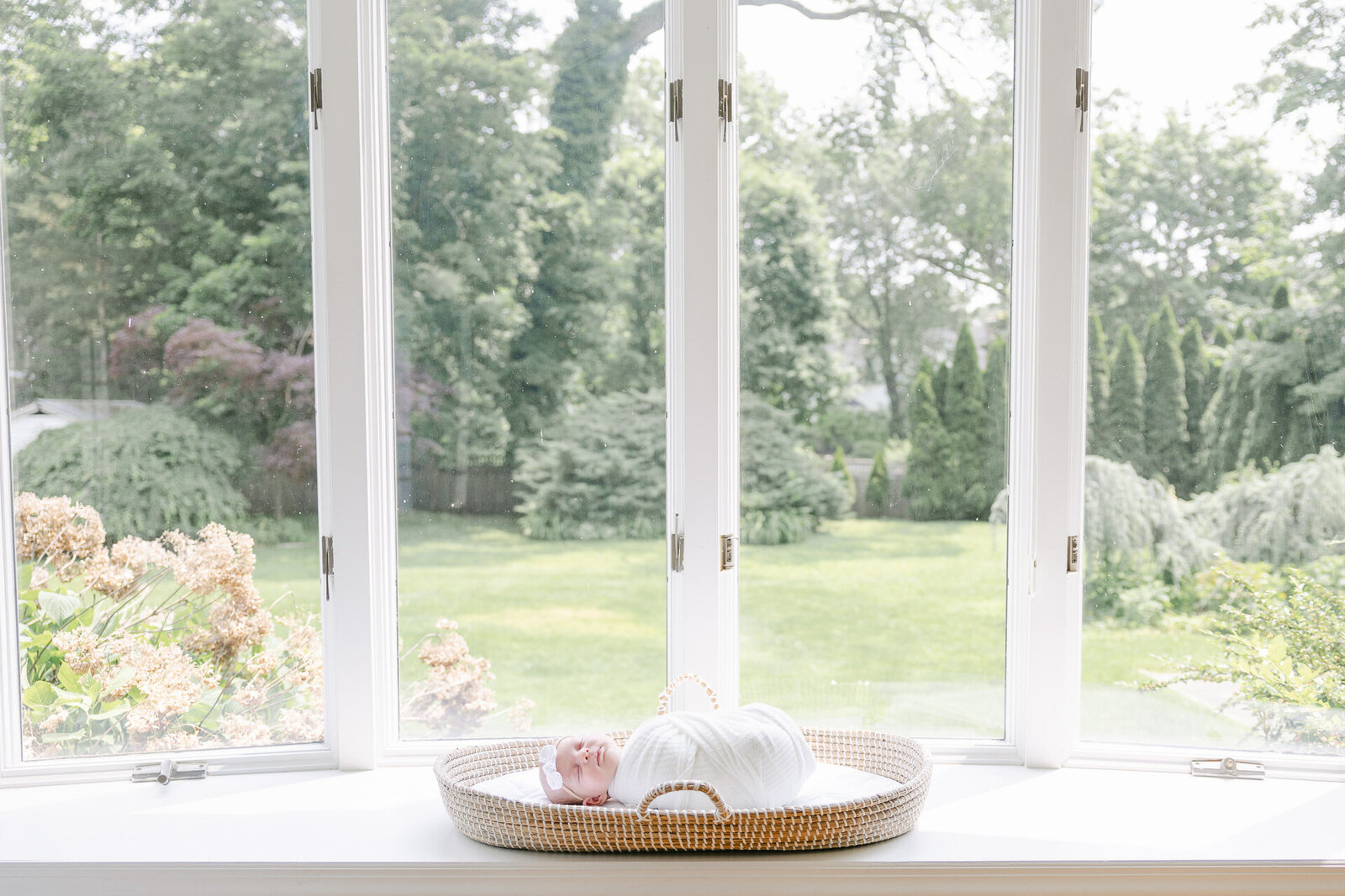 A newborn peacefully swaddled in white rests in a woven basket on a bright windowsill, surrounded by soft natural light and a lush backyard view. A perfect example of what a calm, cozy in-home newborn session can look like in Connecticut. This image highlights the simplicity and beauty of capturing newborn moments right at home—with no studio stress required.