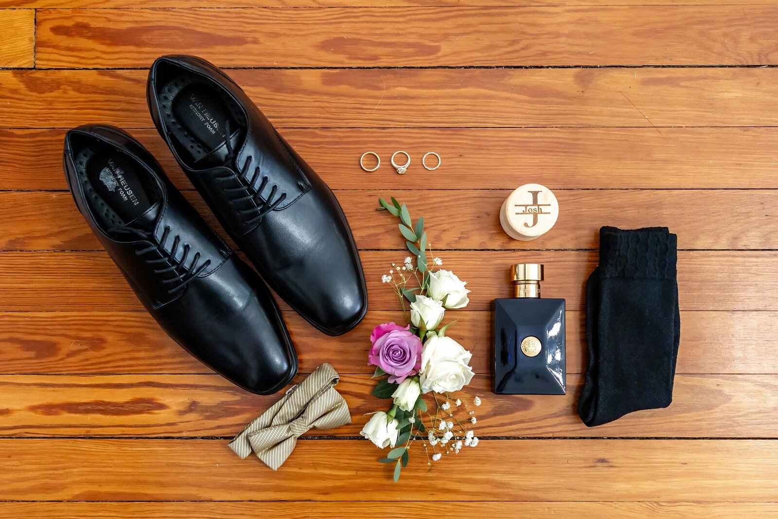 A flat lay of the groom's accessories, including polished black shoes, gold bow tie, cologne, and wedding rings, captured on wooden flooring during a Wrightsville Beach, NC wedding, blending elegance and personal style.