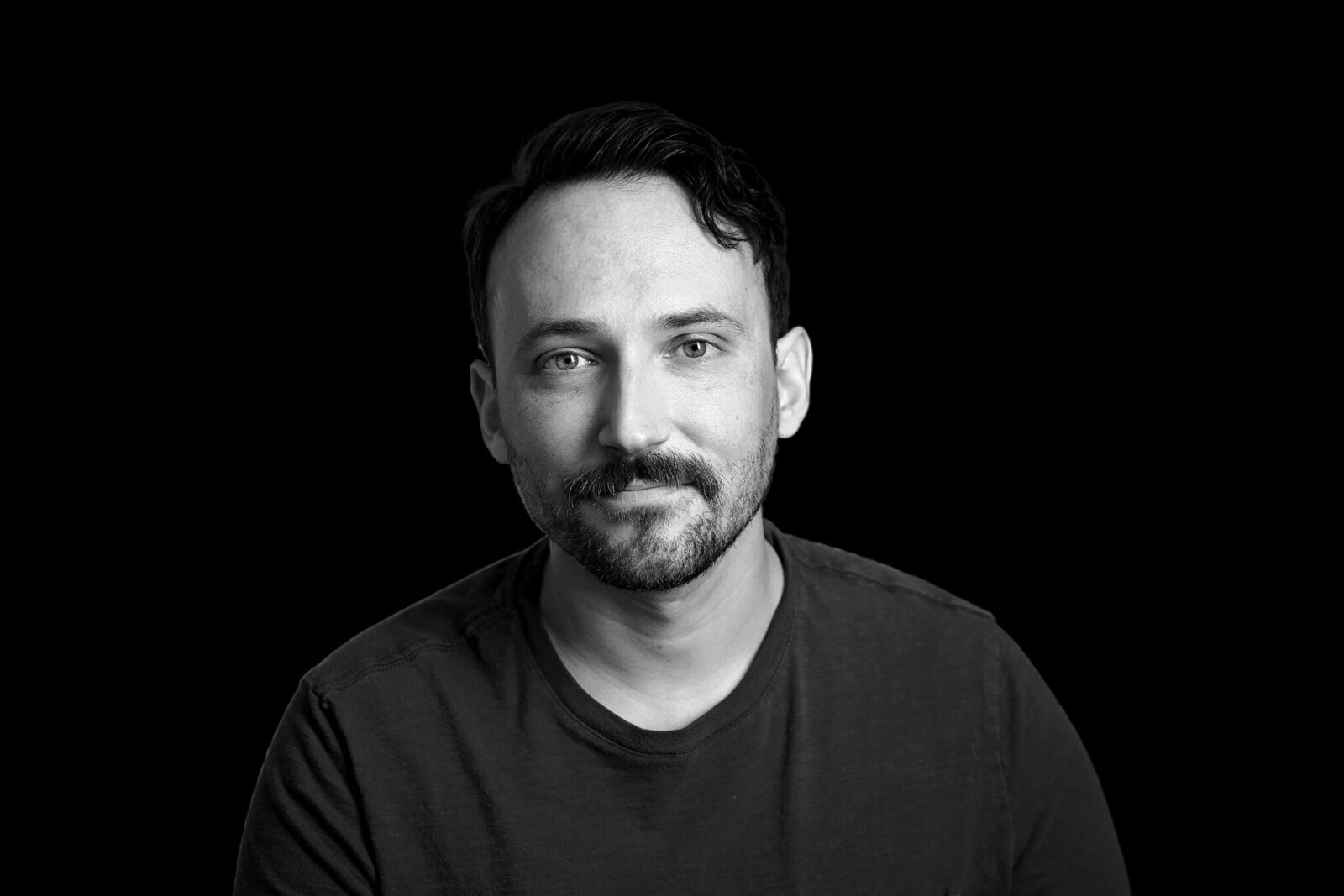 Black and white portrait of a man with short hair and a mustache, wearing a plain t-shirt, looking directly at the camera against a solid black background—ideal for professional band photos Jacksonville.