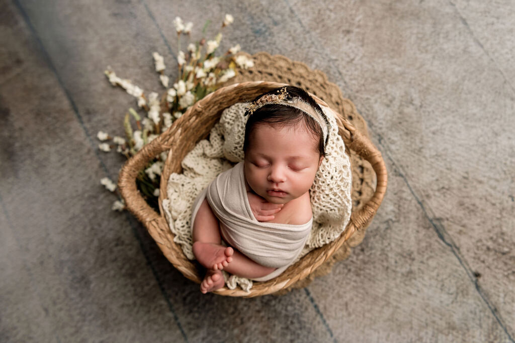 Sleeping newborn nestled in woven basket with knitted blanket and dried flowers on wooden floor