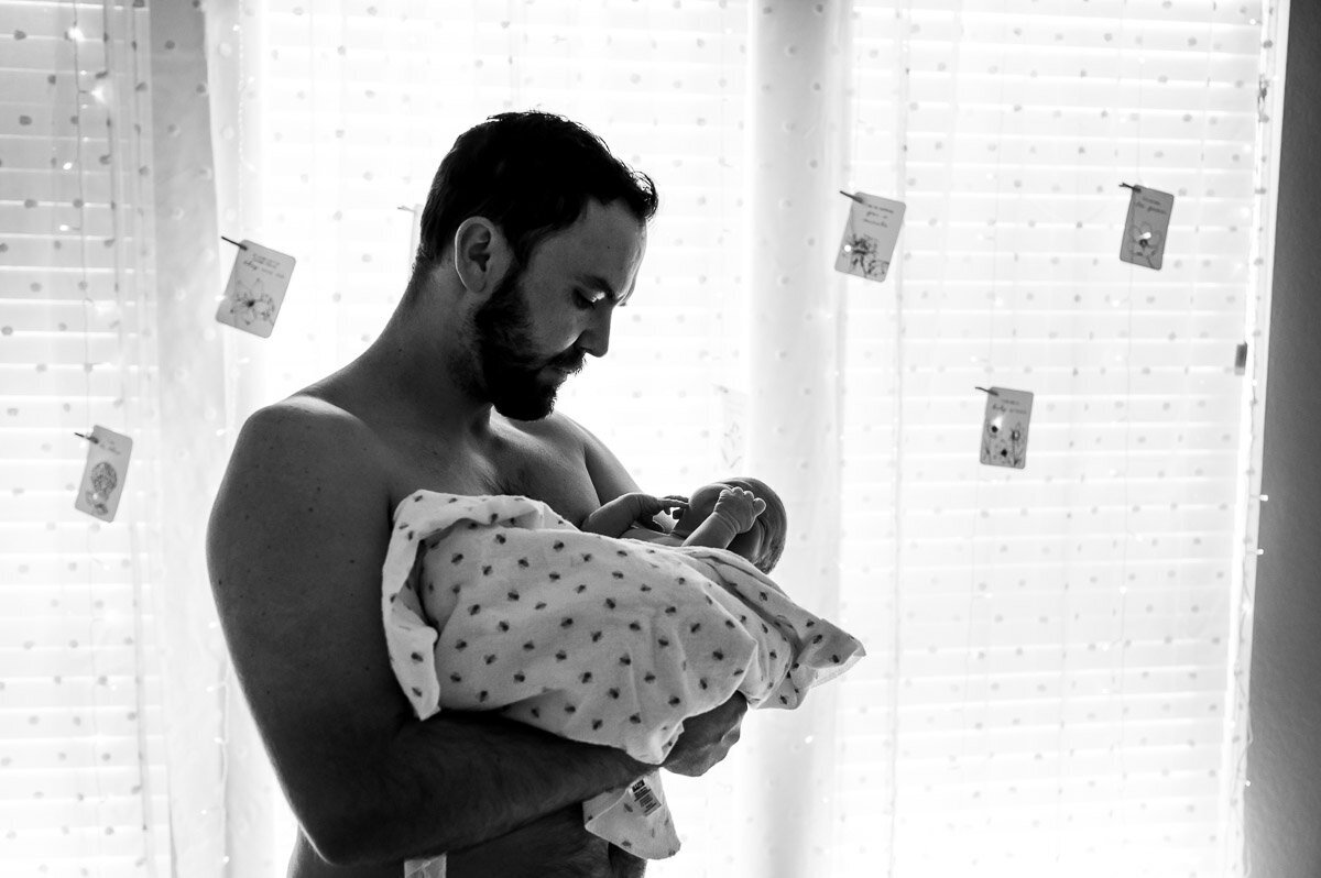 dad stands by window holding newborn at home