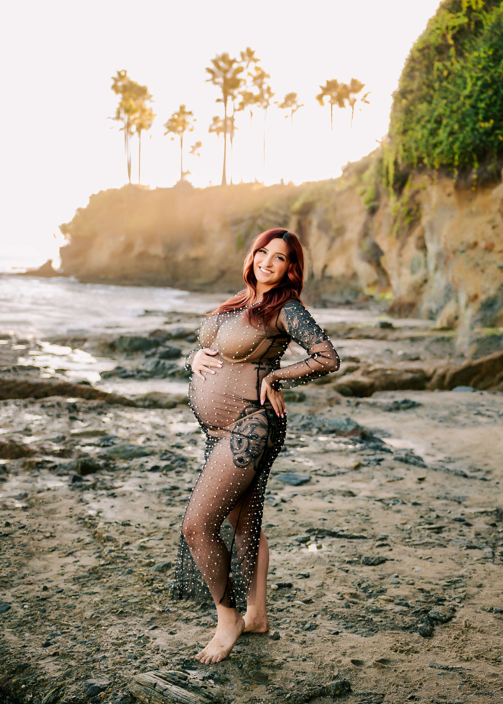 Expectant mom posed on rocks in laguna beach wearing peal sheer dress by Ashley Nicole.