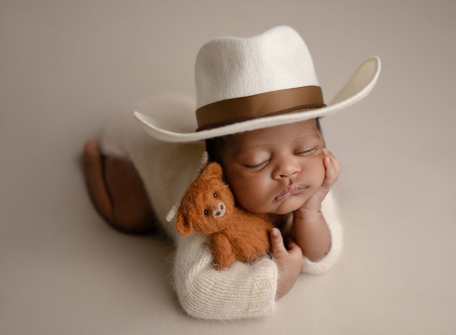 Baby on tummy with hand under chin and wearing a cowboy hat and holding felted highland cow. Newborn Photography session photographed at Jenni Nikki's photo studio in Marietta, GA.