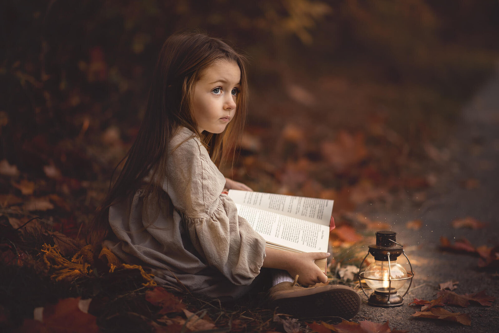 little girl sitting on the ground with a book in her lap and lantern by her side