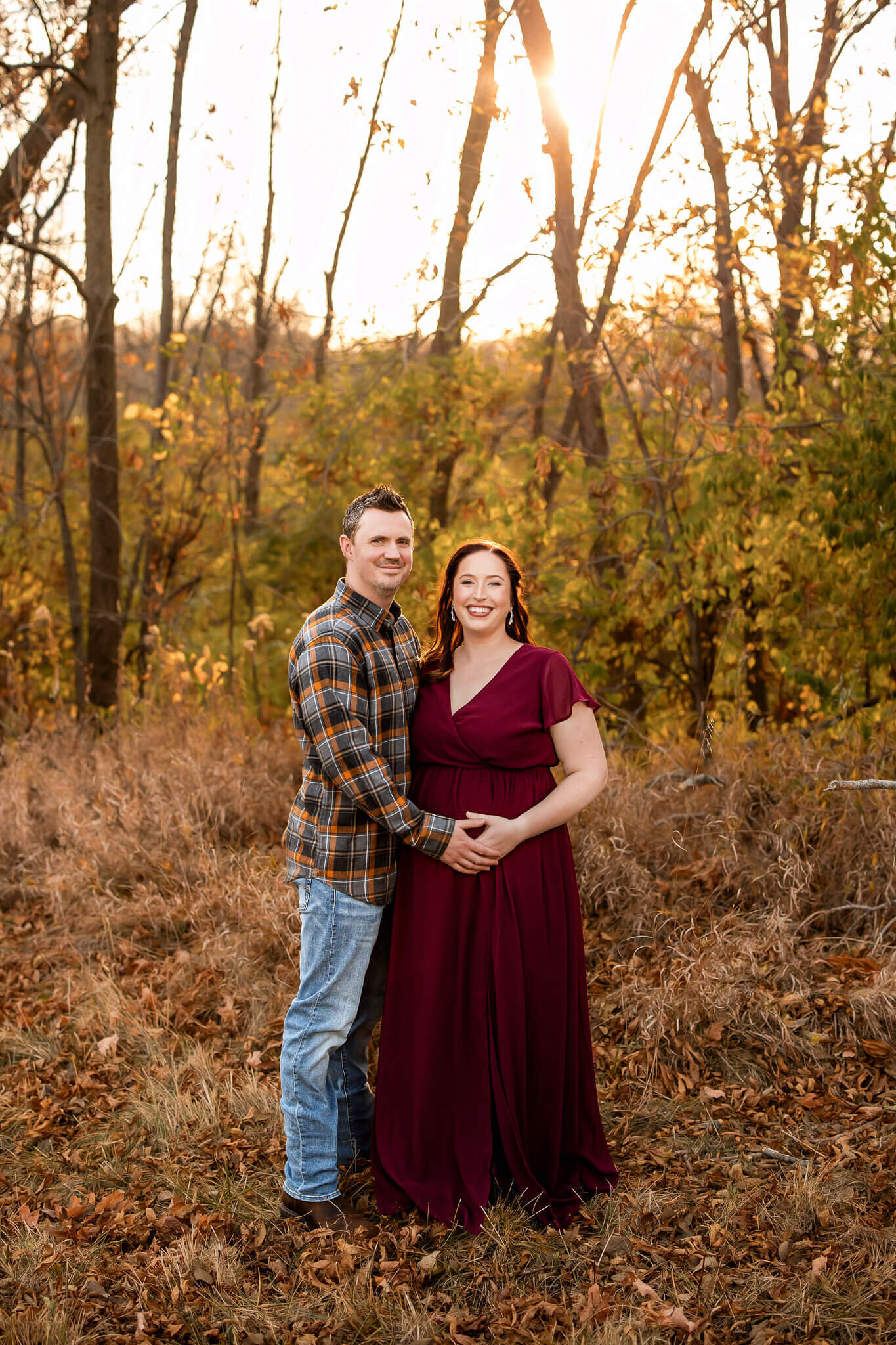 A smiling couple stands together outdoors in autumn. The man wears a plaid shirt and jeans, while the woman in a flowing burgundy dress cradles her belly. Sunlight filters through golden leaves—perfect for Bloomington maternity photographer maternity portraits.