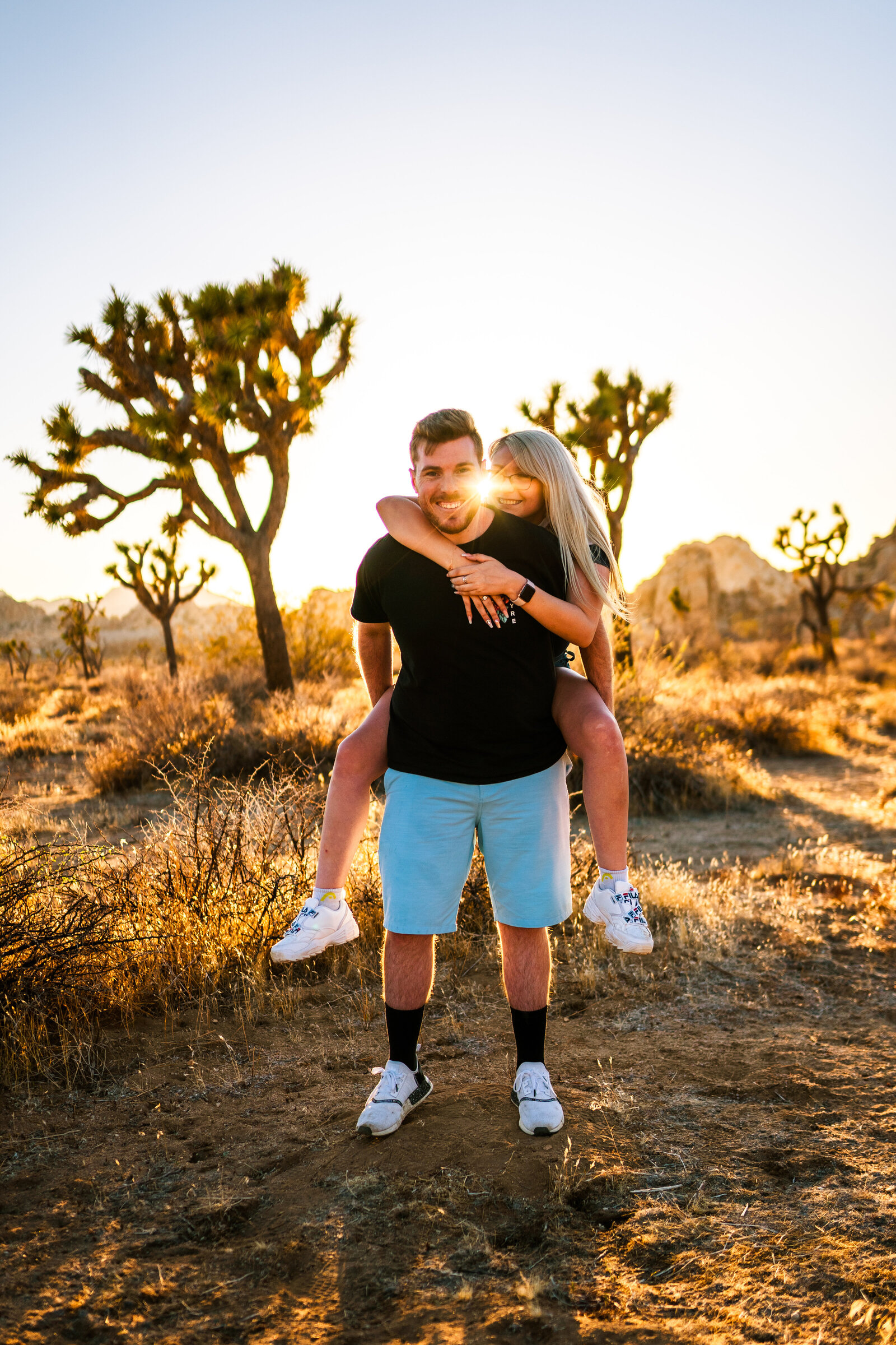 Man carrying woman on his back, with Joshua trees behind them and the sun setting.