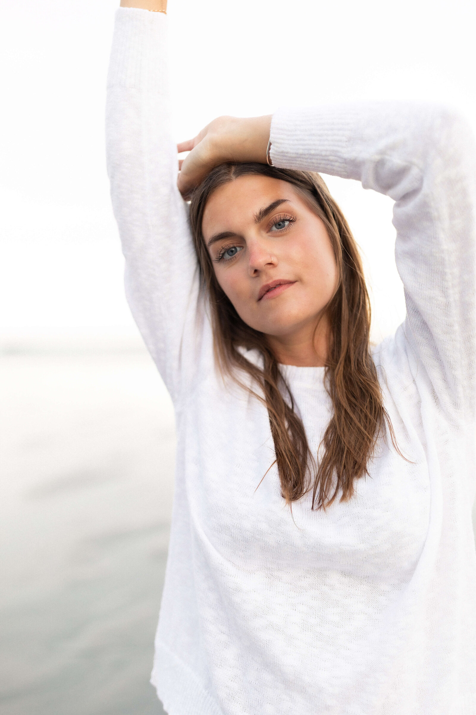 brunette woman wearing long sleeve white top with hands over her head posing for camera