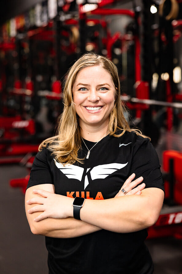 Girl with Kula shirt on folds arms and poses in front of red squat machines in a gym.
