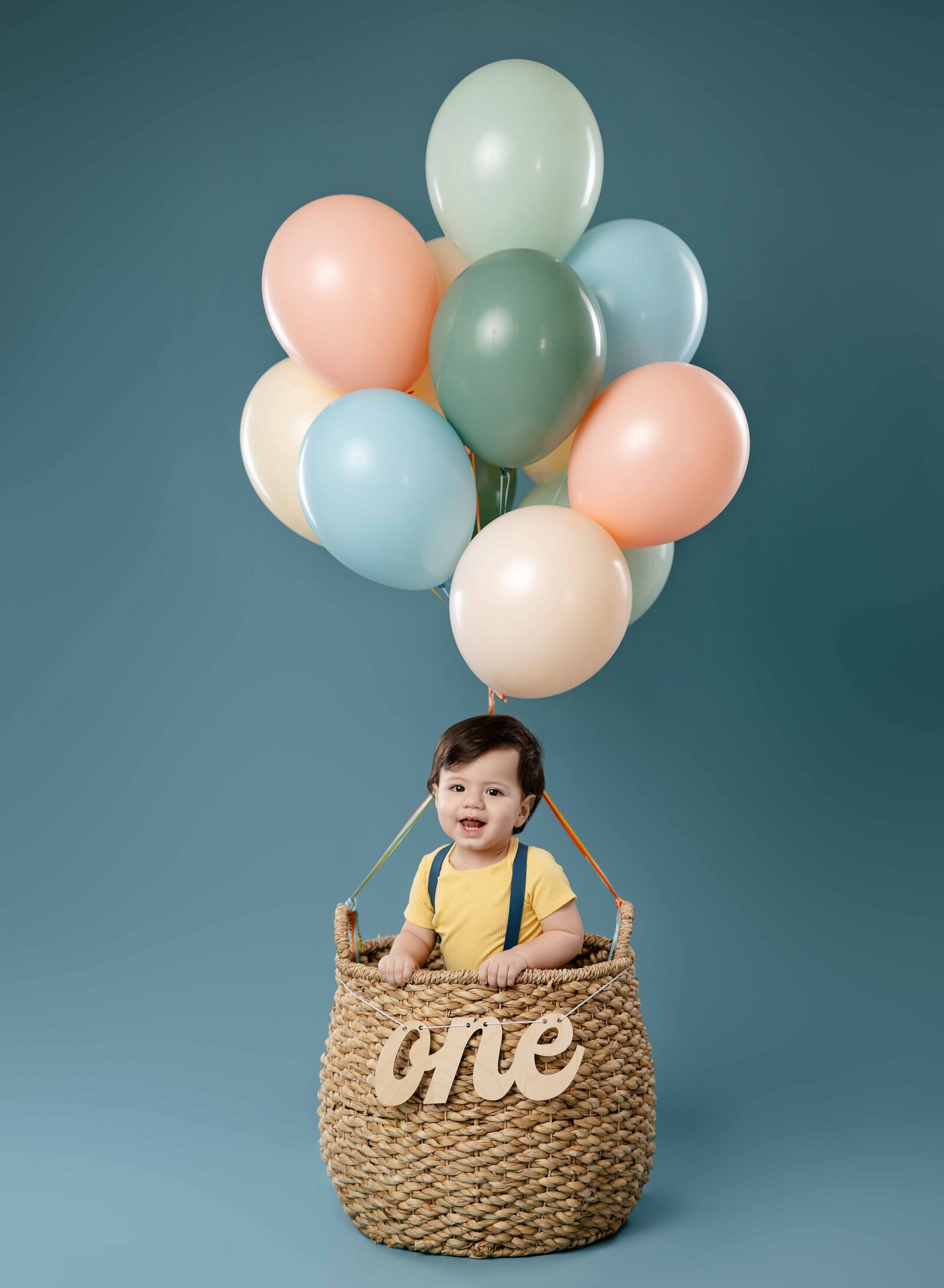 1 year old in balloon basket with pastel boy color balloons and smiling at the camera.