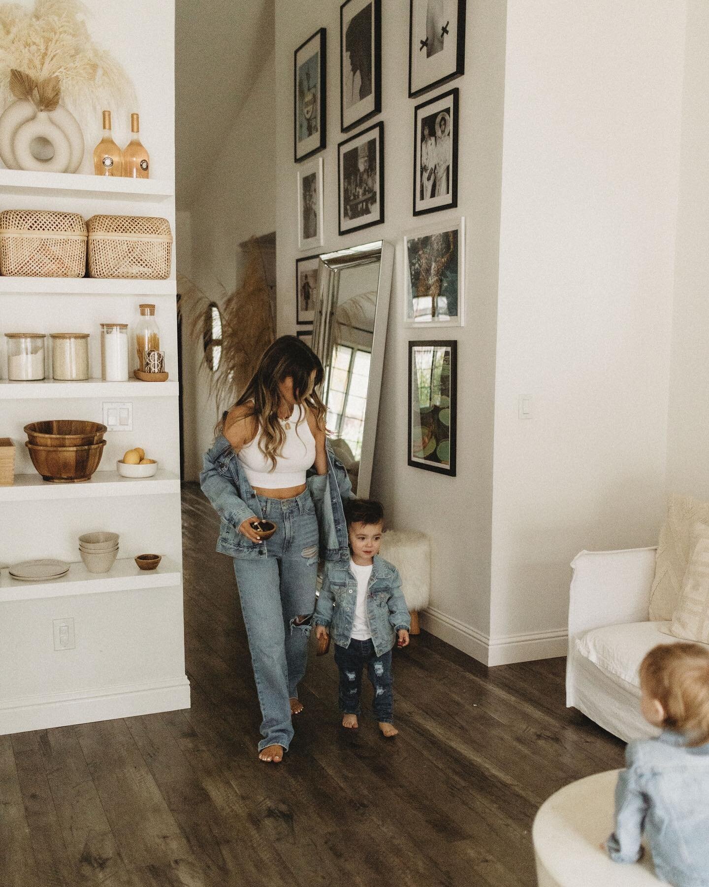 A woman in denim holding hands with a toddler in a stylish home. they walk towards a sitting baby, surrounded by shelves, framed photos, and cozy decor.