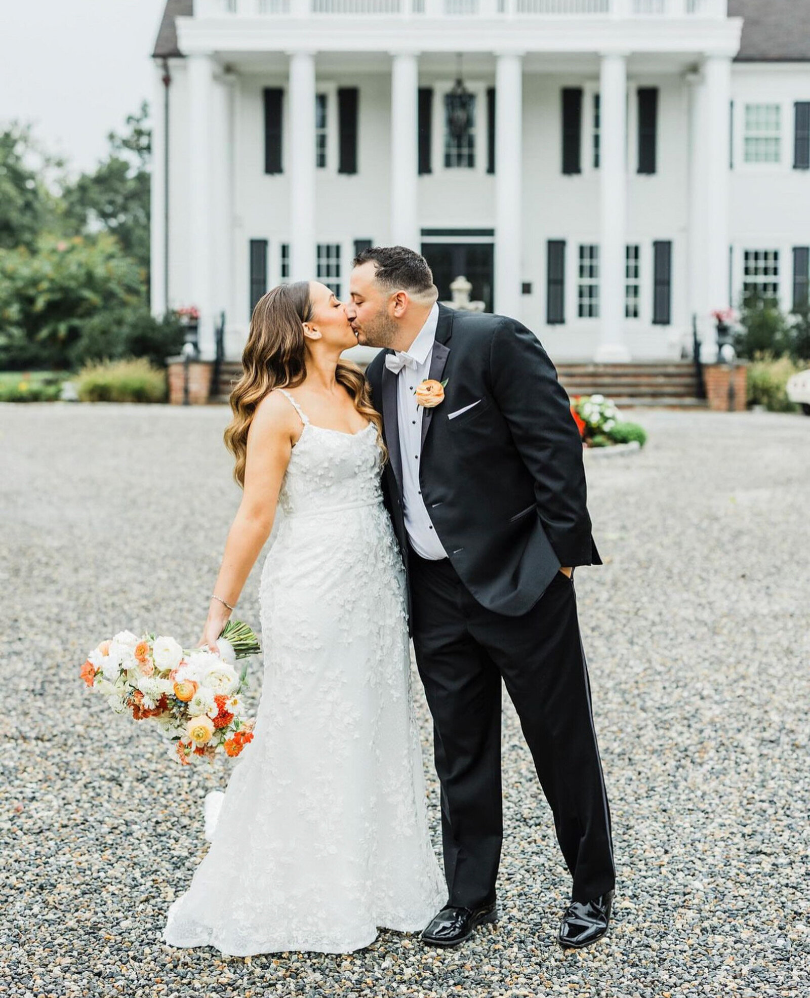 bride, in a white lace wedding dress, holds a bouquet of flowers, while the groom, in a black suit with a white shirt and orange boutonniere, leans in for the kiss.