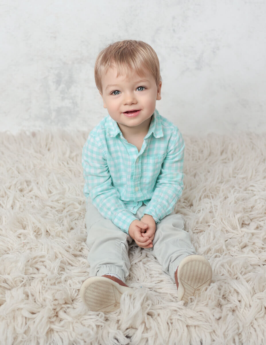 A joyful two-year-old child sitting on a soft white rug during a milestone photography session in a studio in Rochester, NY. The child is smiling, surrounded by a neutral backdrop that enhance the atmosphere of the event.