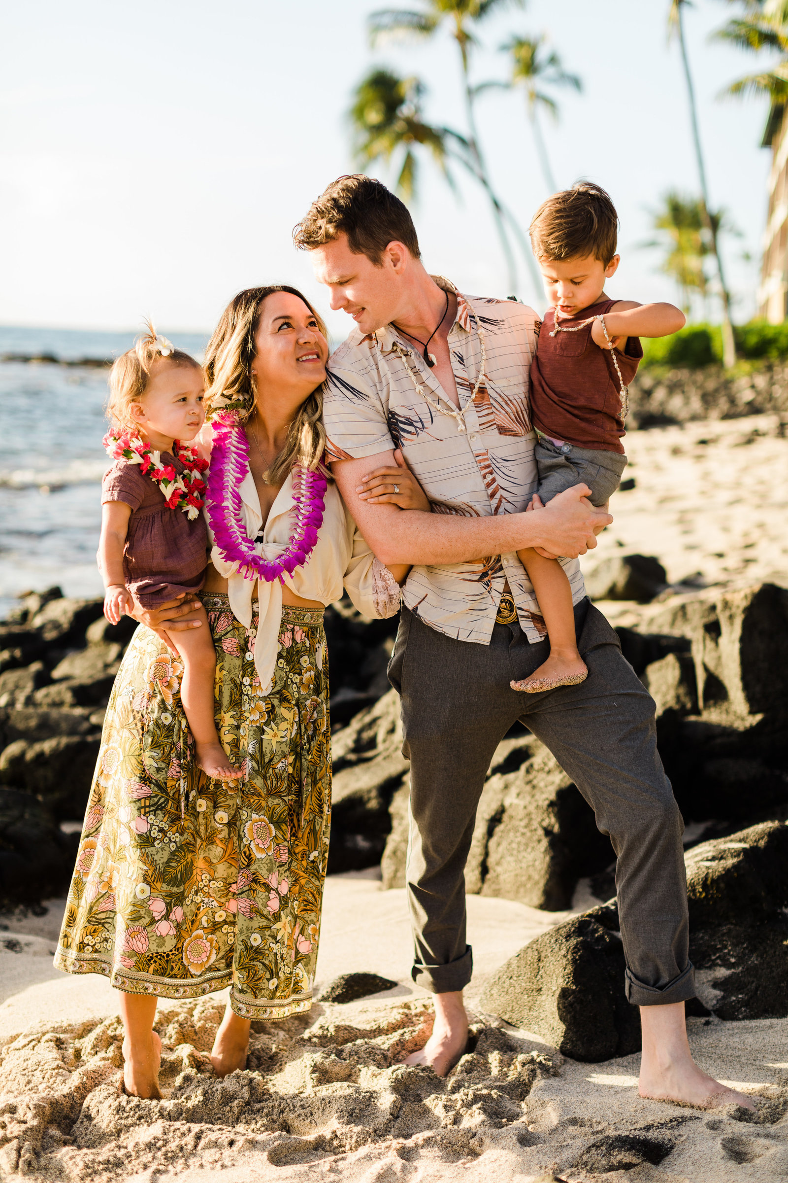 Young couple hold their two young kids on a beach in Hawaii, surrounded by lava rocks and palm trees while wearing leis