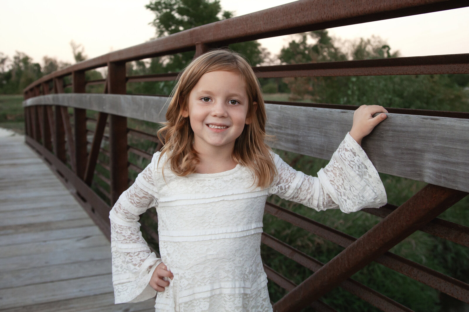 Girl in cream dress with one hand on bridge railing, smiling.