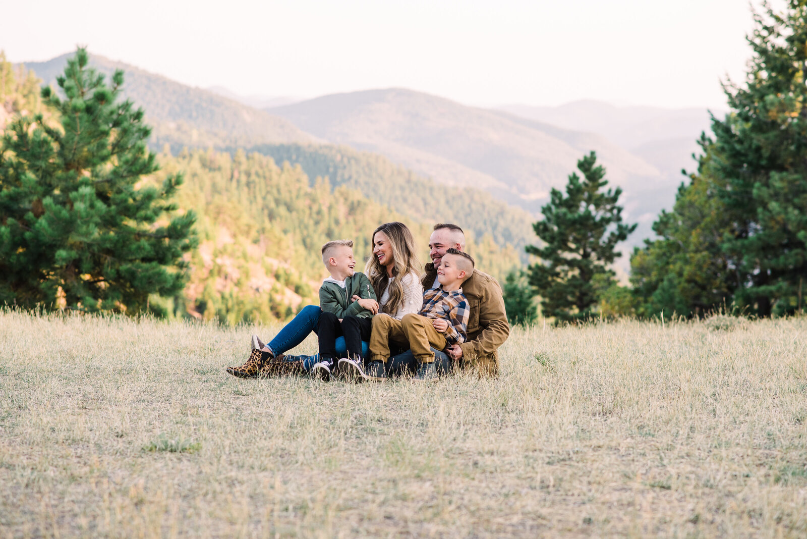 A family sits in the grass of a big field with a large view of the mountains behind them and a few sparce evergreen trees