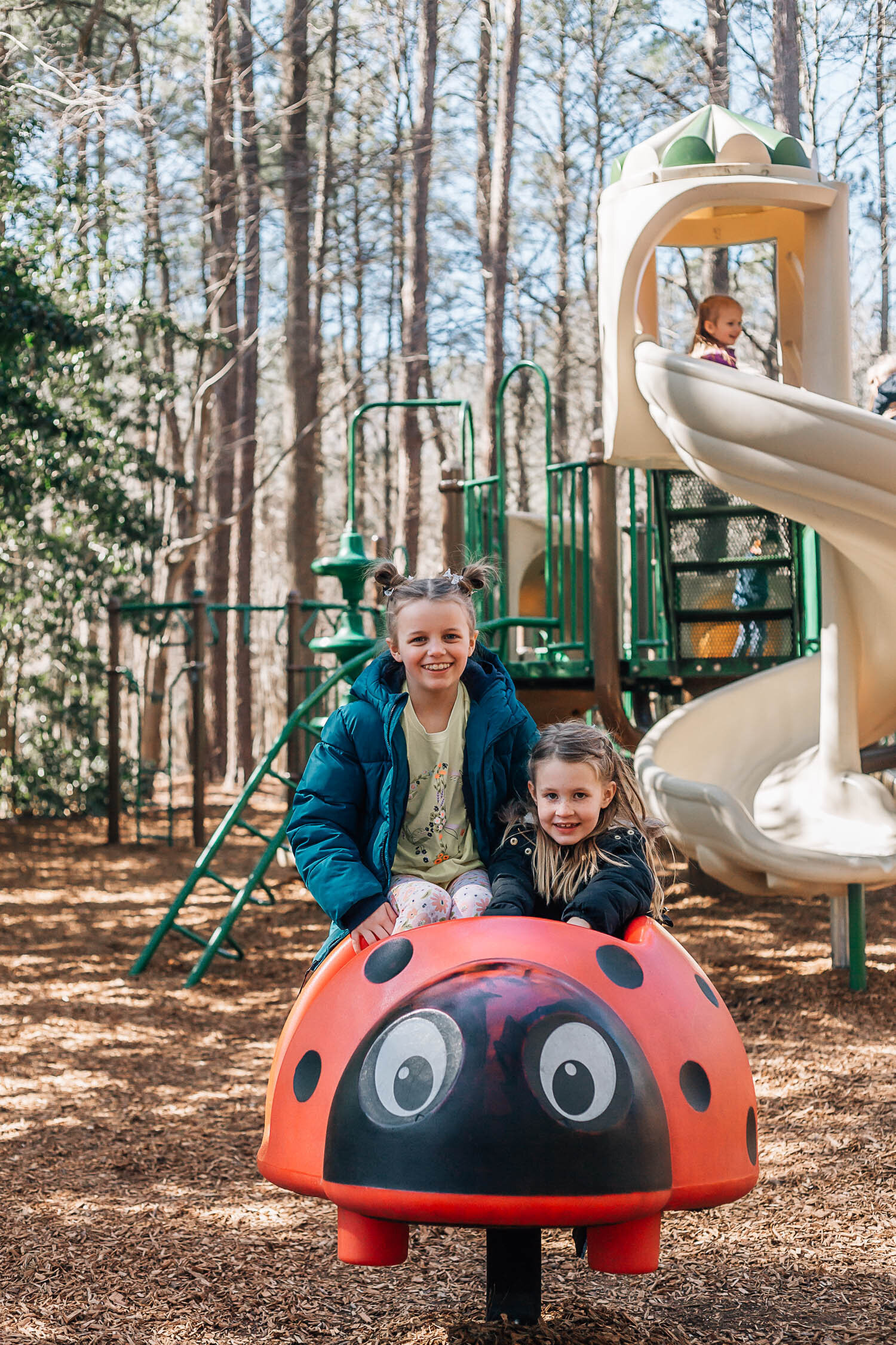 two primary aged sister sitting on a lady bug toy at the play ground and smiling as they play in the fall captured by the best Hampton Roads Family photographer