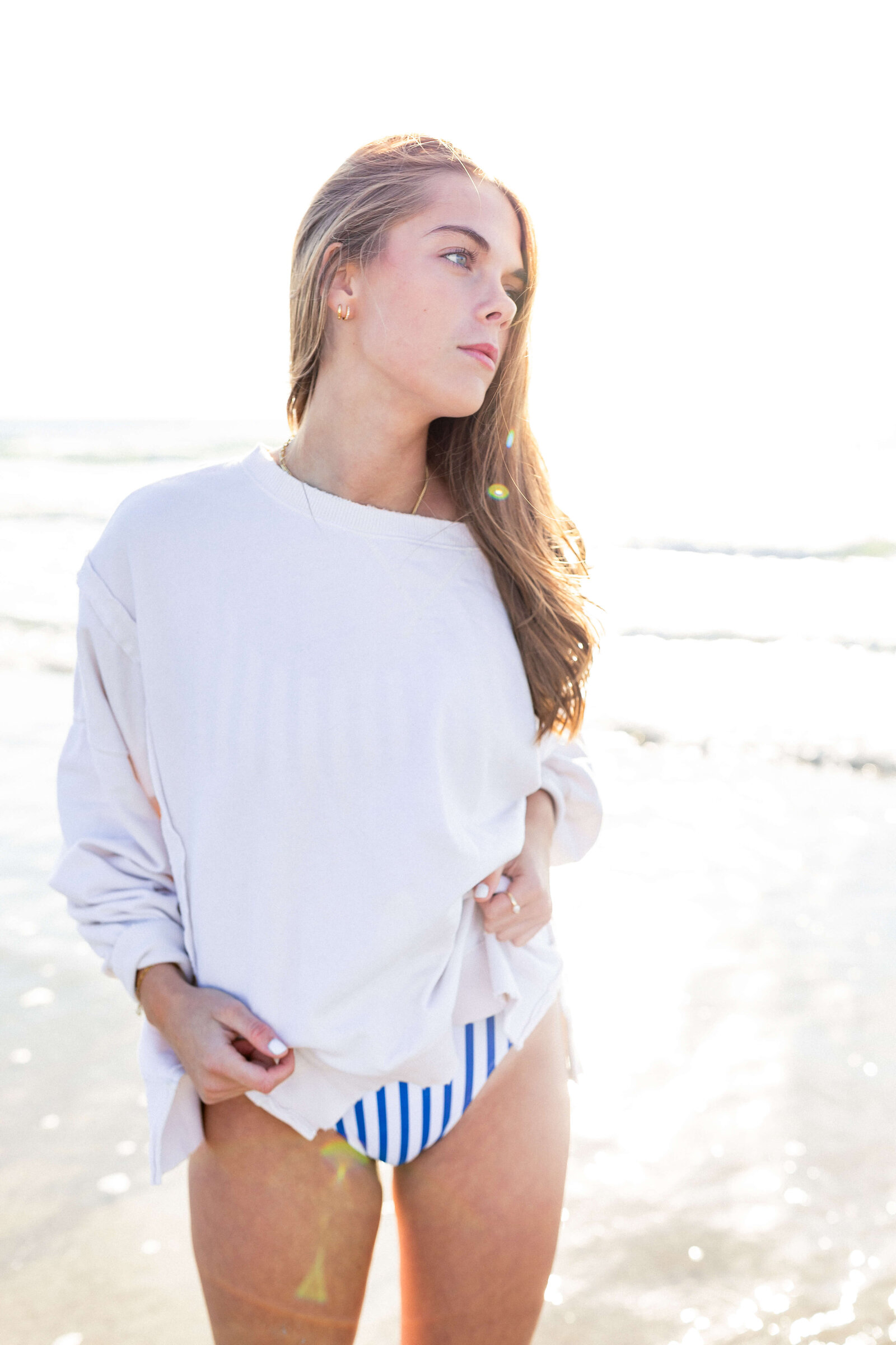 brunette high schooler posing on a beach wearing a white shirt and blue striped swim suit bottoms
