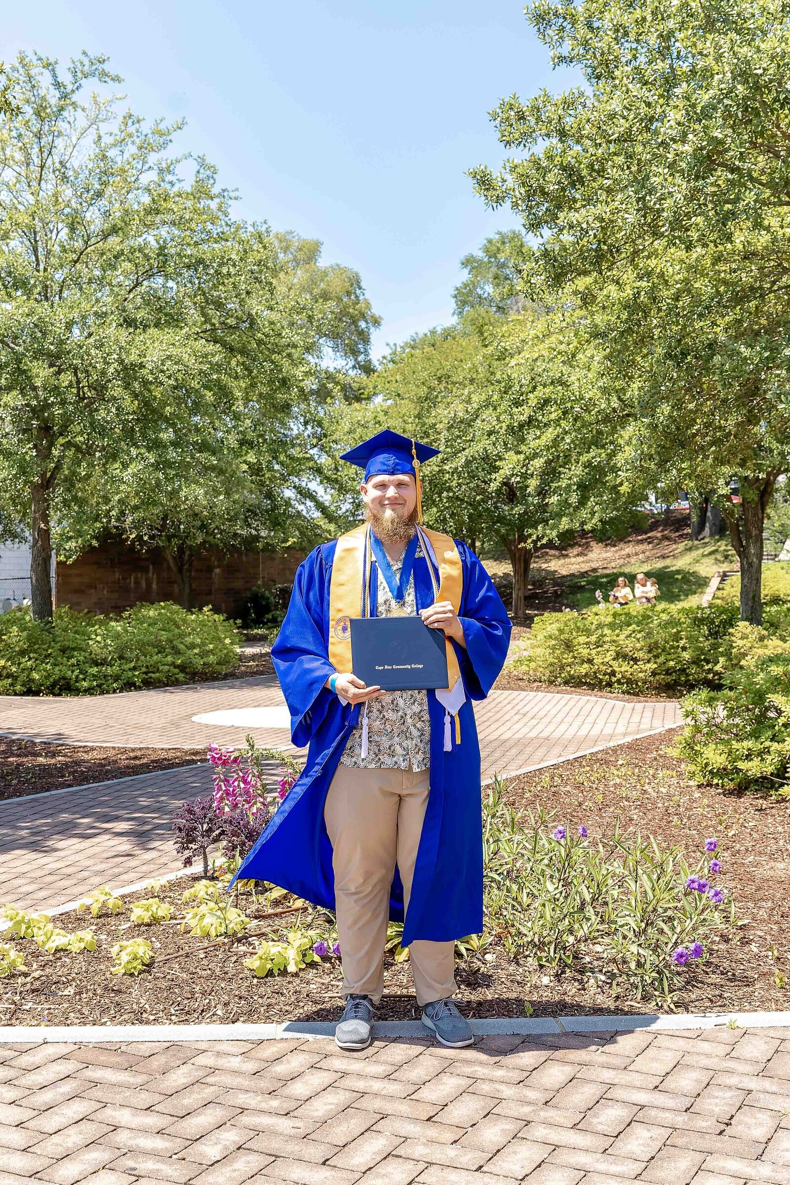 A Cape Fear Community College senior in a blue cap and gown proudly holding a diploma in a garden setting in Wilmington, North Carolina. This image celebrates academic achievement, perfect for capturing professional and joyful senior portraits.