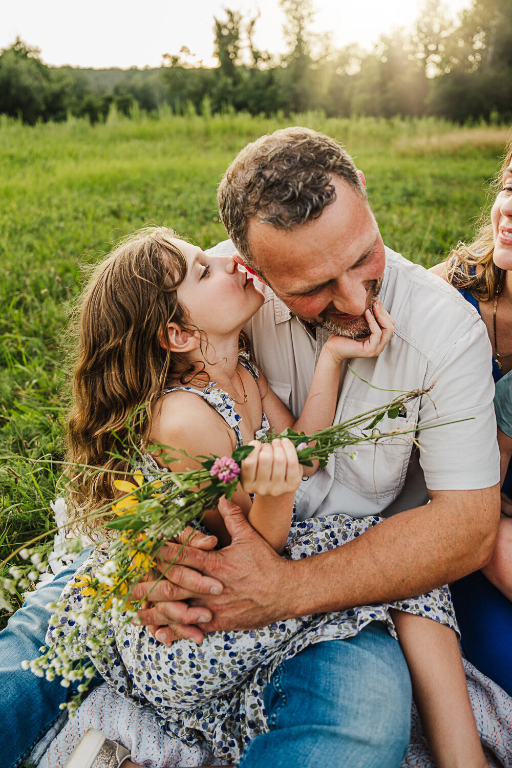 girl sits on her fathers lap and whispers to him while holding a bouquet of wildflowers