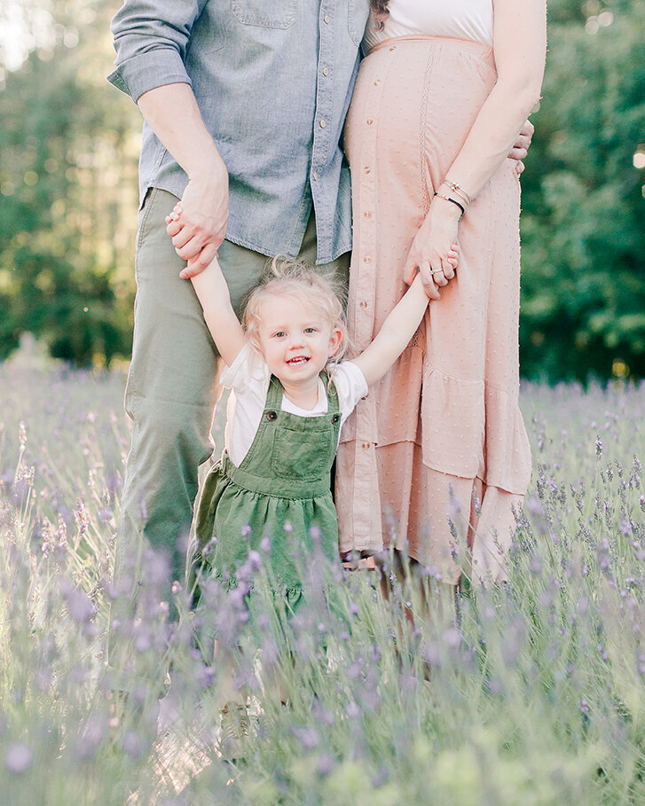 Three year old in green overall dress holding her parents hands in a lavender field by NH newborn photographer Fieldstone Studio.