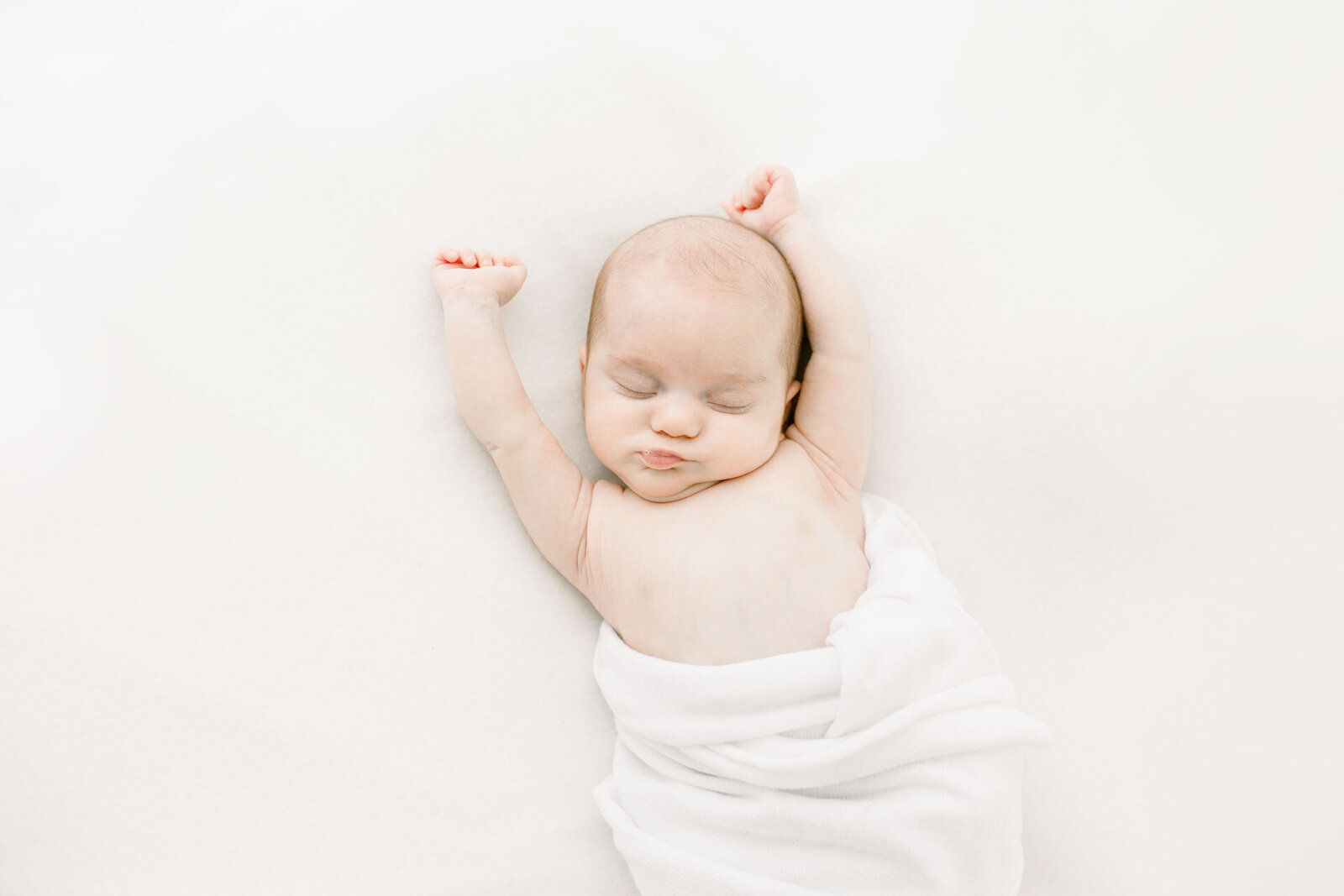 Neugeborenenshooting: Schlafendes Baby mit ausgetreckten Armen in weiße Decke gewickelt.
