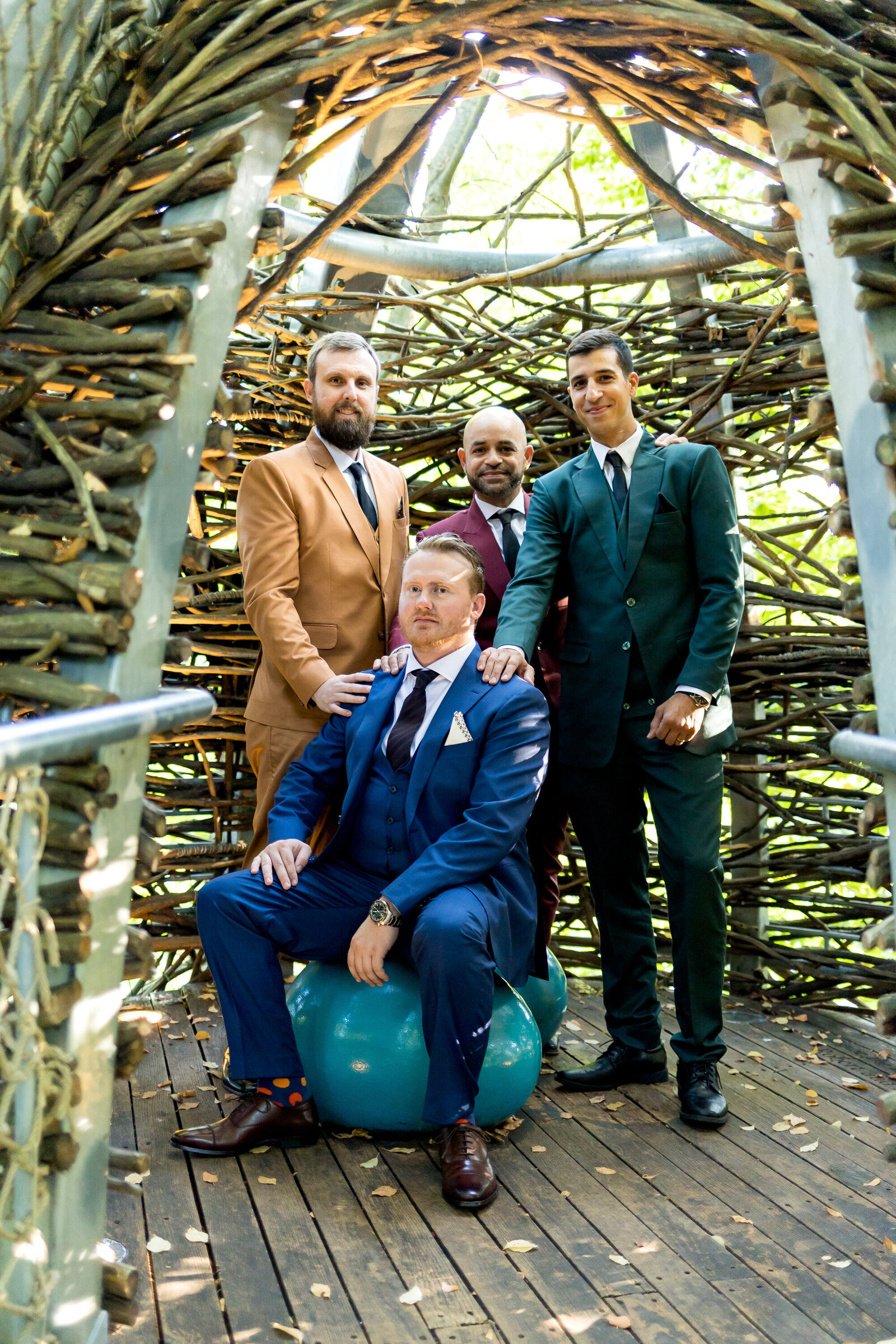 4 groomsmen posing in arboretum. The groomsmen are wearing fall coloring suits (red, blue, orange, green, and yellow).