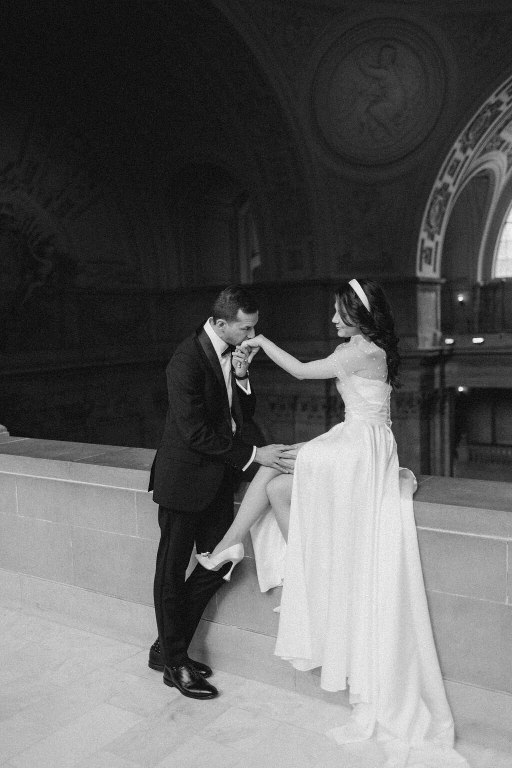 A black and white photo of a bride and groom on a grand staircase in San Francisco City Hall, with the groom kissing the bride's hand. The setting features elegant architectural details.