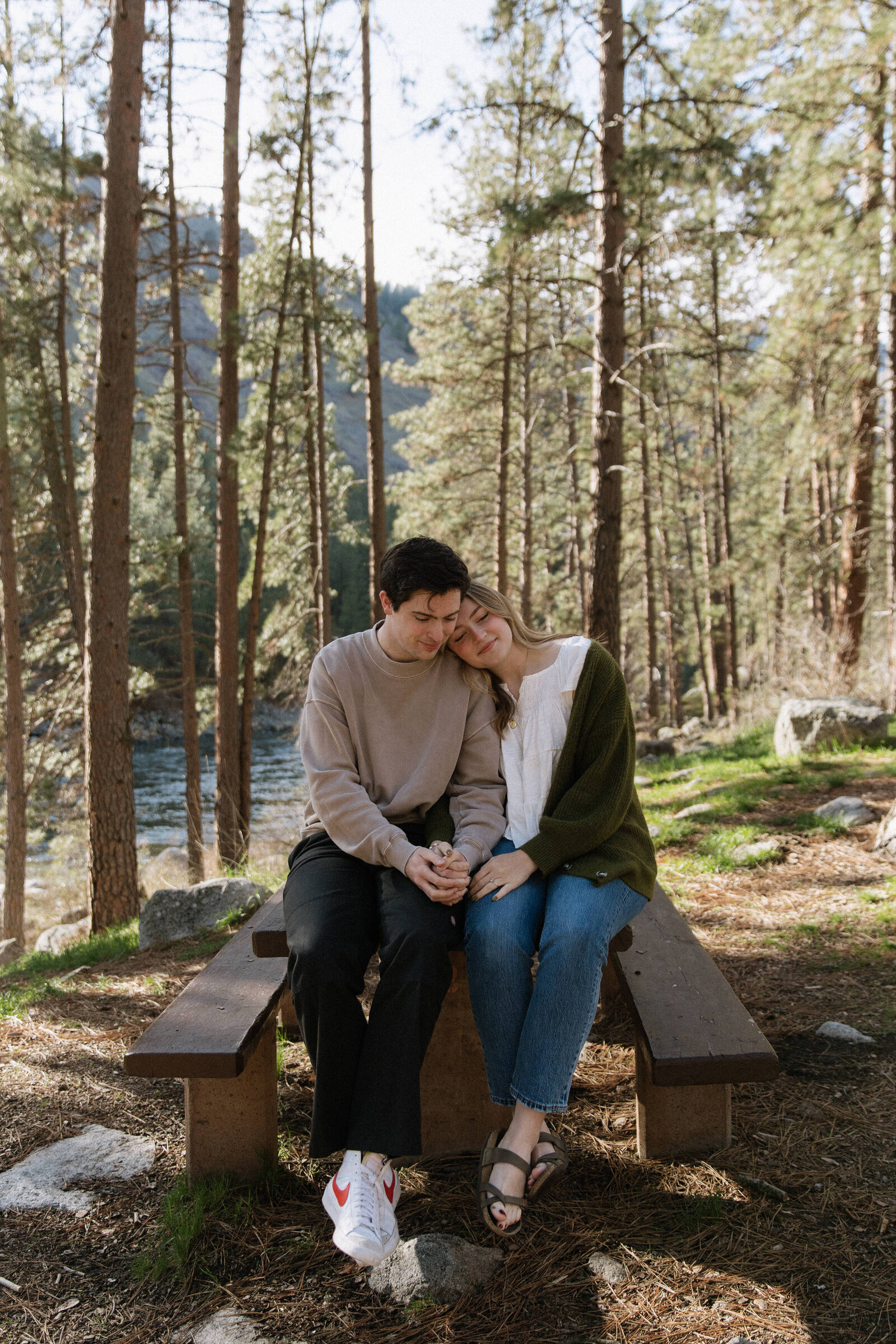 Sweet engagement photo of couple cuddled on rustic picnic table by the water
