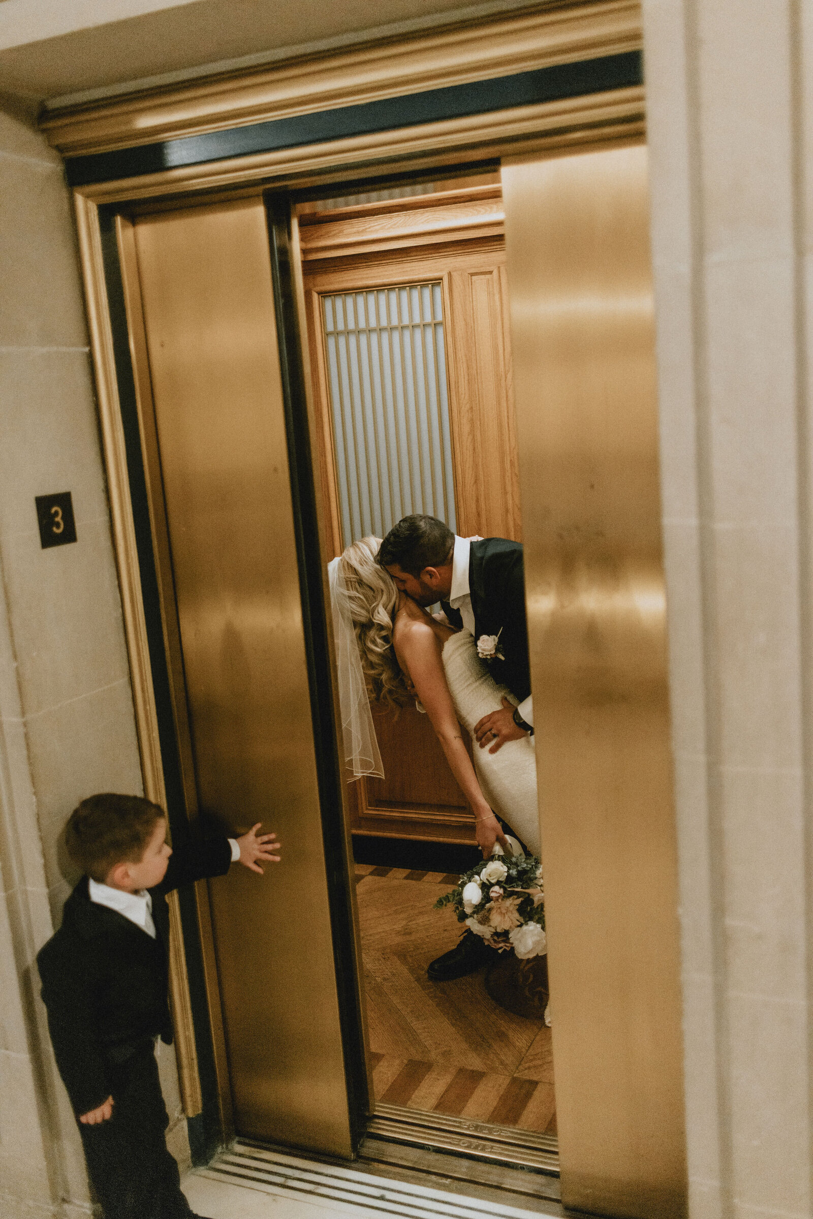 A bride and groom share a kiss in an elevator during a photoshoot, holding a floral bouquet, as a young boy watches them, all inside a vintage elevator with wood paneling in San Francisco City Hall.