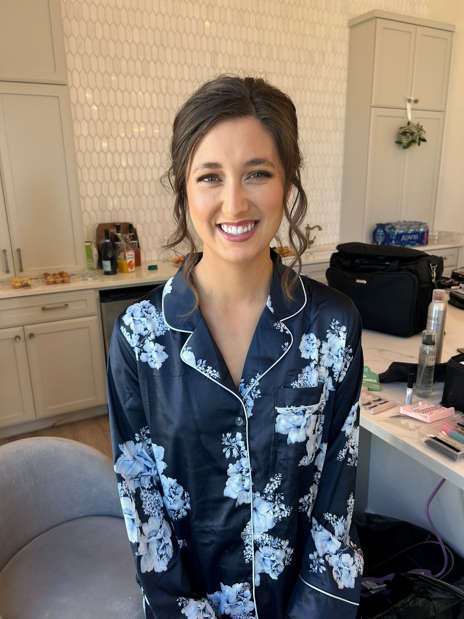 A woman in floral pajamas smiles warmly in a kitchen setting, surrounded by makeup items. Her relaxed demeanor hints at the wedding beauty consultation she enjoyed, bringing vibrant life to bridal hair and makeup processes just like this cozy moment suggests.