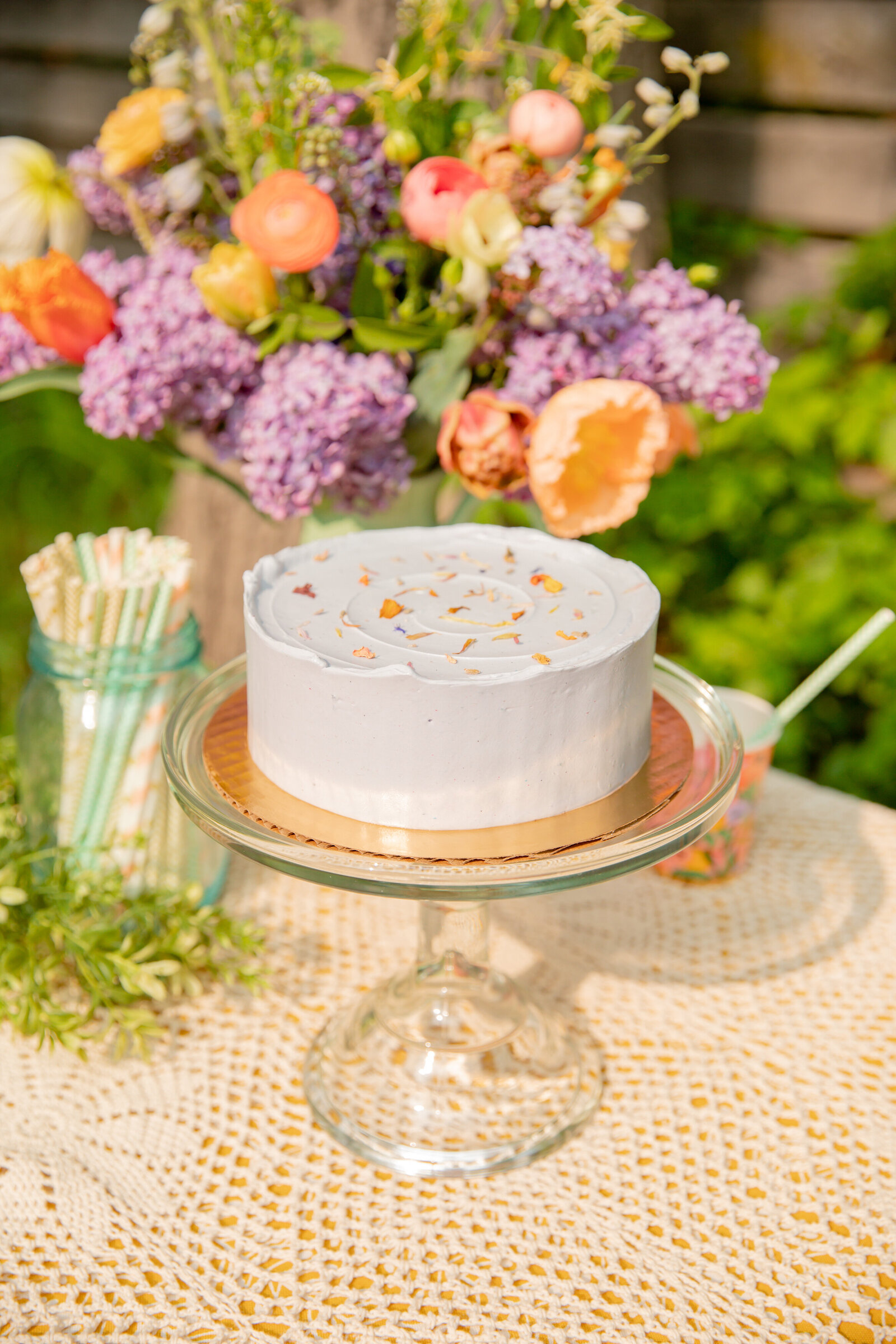 lavendar sweet heather anne cake on class stand with flowers in the background during styled shoot