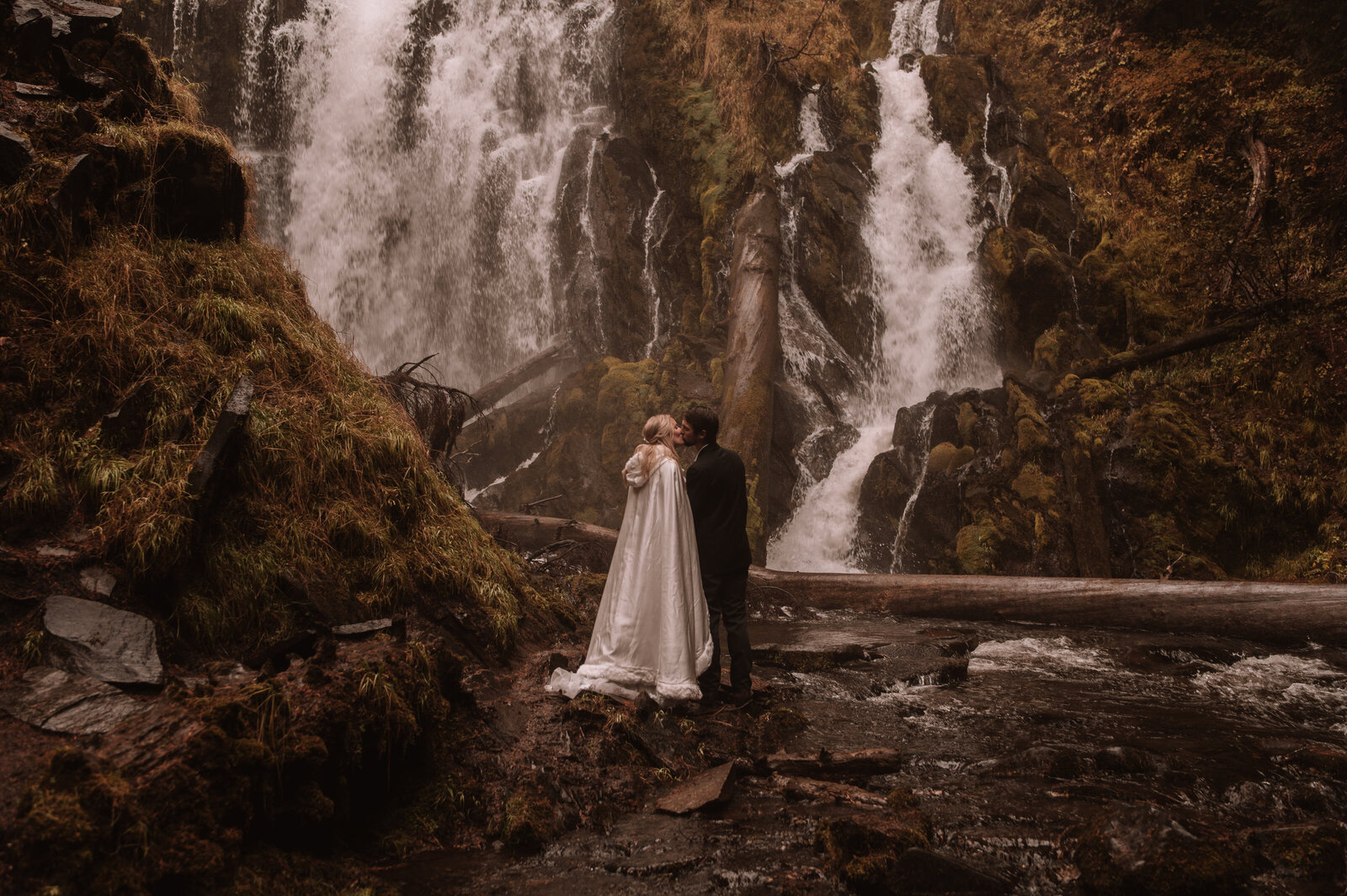 A bride and groom share their first kiss as husband and wife underneath a huge waterfall with fallen logs and green moss surrounding them. They are standing on the bank of the river at the bottom of the waterfall with mist visible in the air.