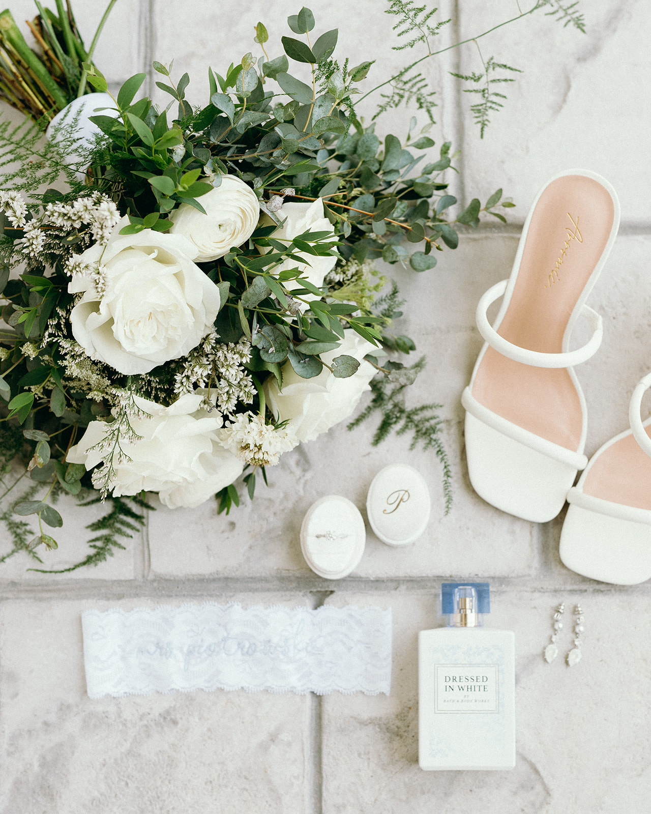 Editorial flat lay of bridal accessories including rings, bouquet, wedding shoes, wedding earrings, and perfume.