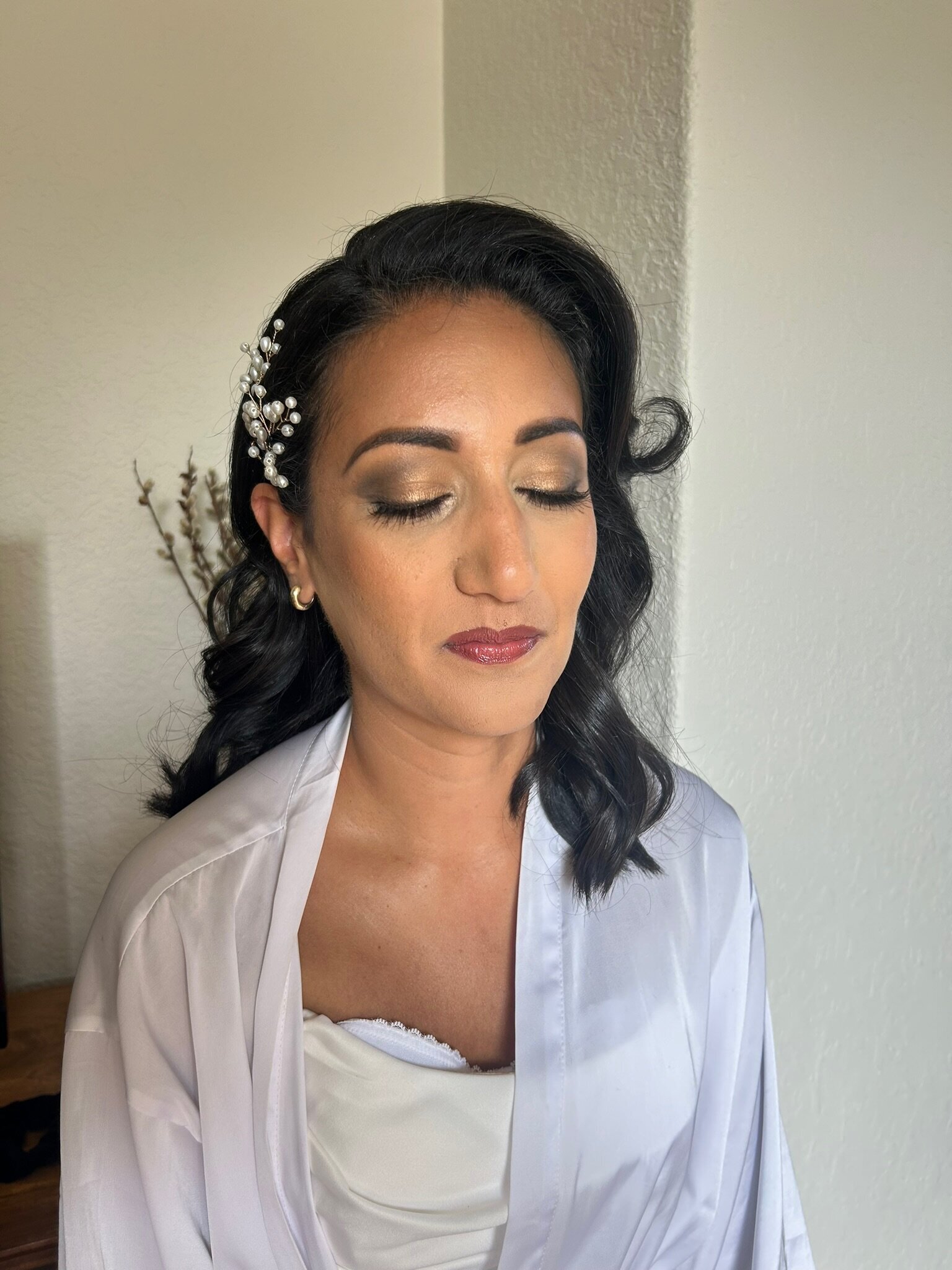 Woman in a white robe with eyes closed, showcasing natural glam wedding makeup and the best wedding hair accessories, including a pearl hair accessory, against a light wall background.
