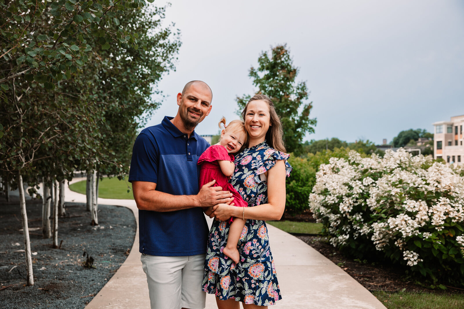 Happy parents stand in a park sidewalk with flowers holding their toddler daughter