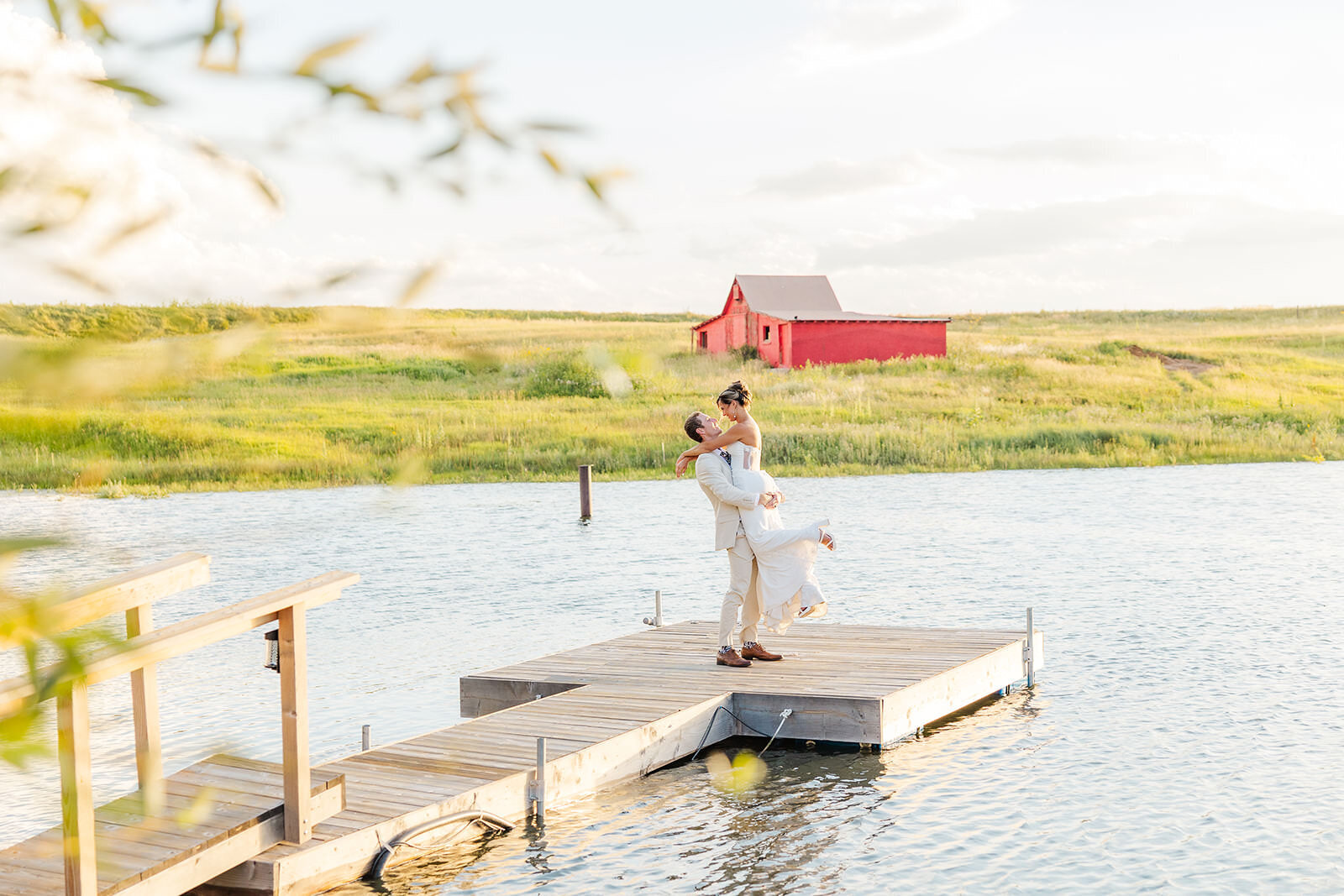 Bride and groom celebrating their wedding day with a romantic lift on the dock in Franktown, Colorado, surrounded by picturesque scenery and a charming red barn backdrop.