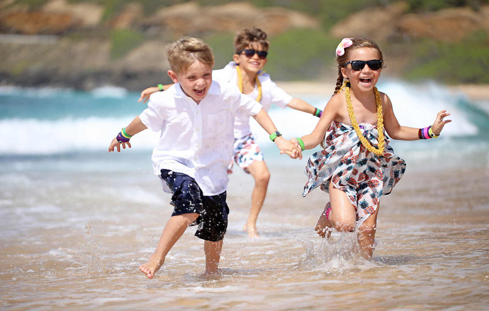 Three children running along the beach in Oahu with ocean water splashing on them for a family portrait photo session
