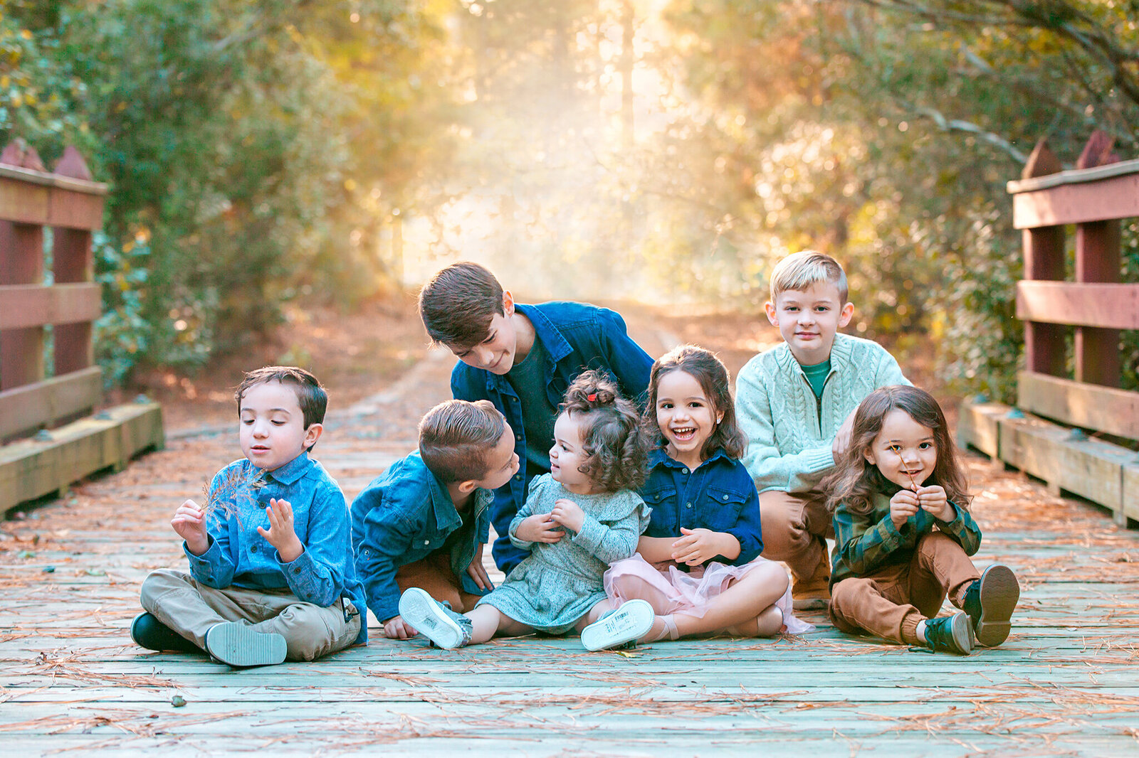 Extended family photos with cousin sitting together on a bridge in the woods as the light shines through the trees in the distance captured by Family photographer Hampton Roads va