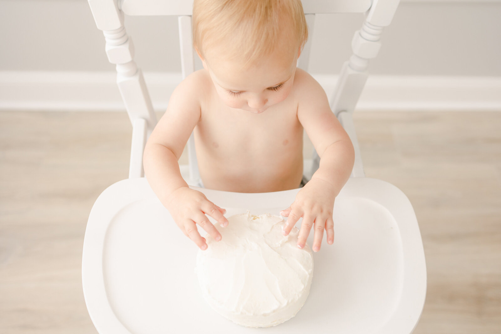 Little boy sitting in his high chair lightly touching his white birthday cake