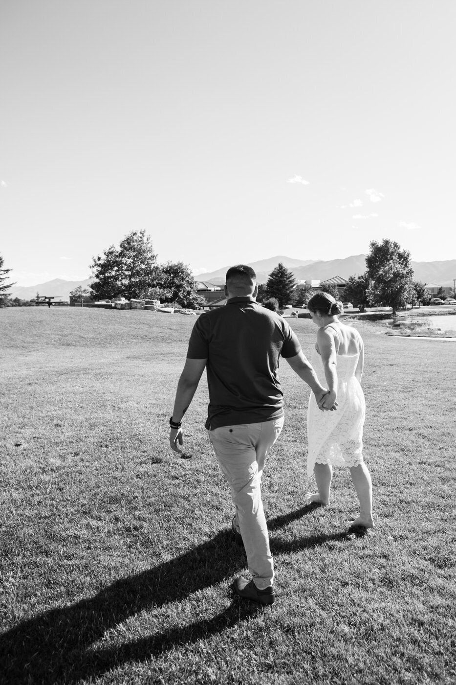 An engaged couple walks away from the camera hand in hand in a grassy field