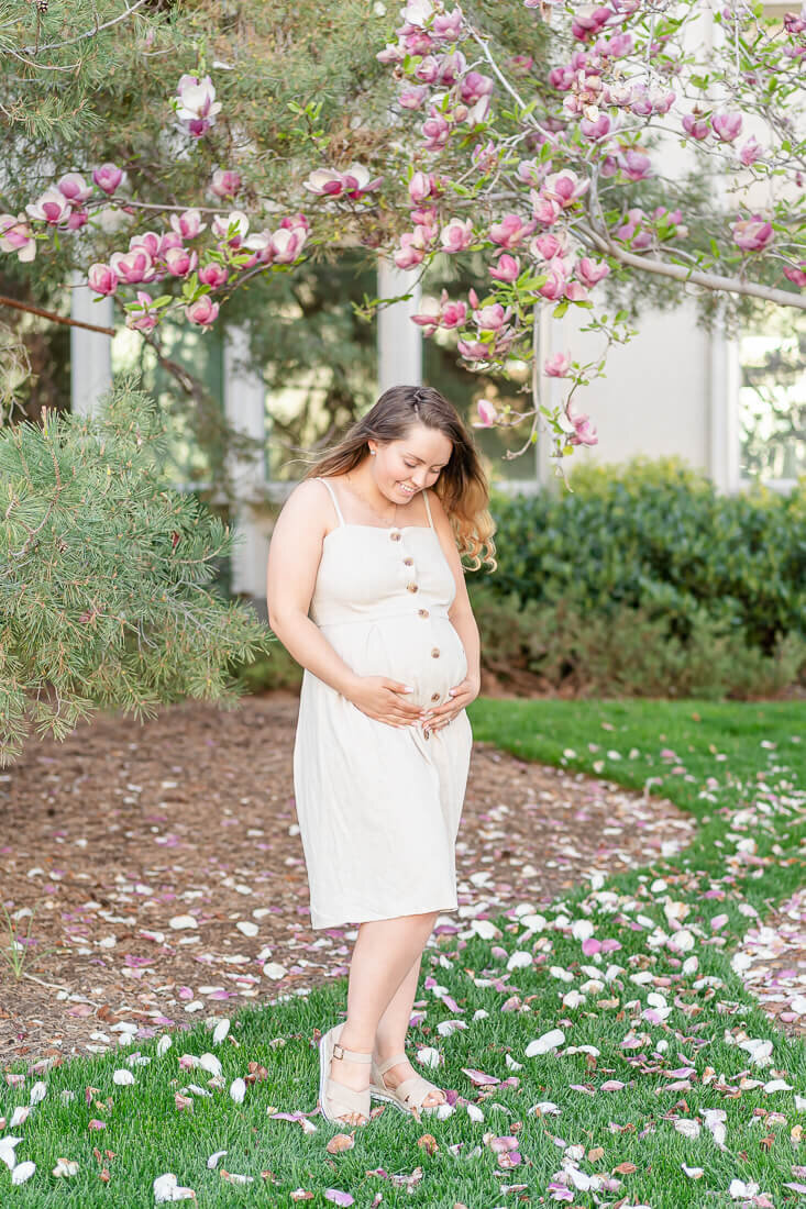 A pregnant woman wearing a cream colored sundress holds her belly while she stands in the grass of the grounds of BYU in the spring. Photo taken by Utah maternity photographer Melissa Woodruff Photography
