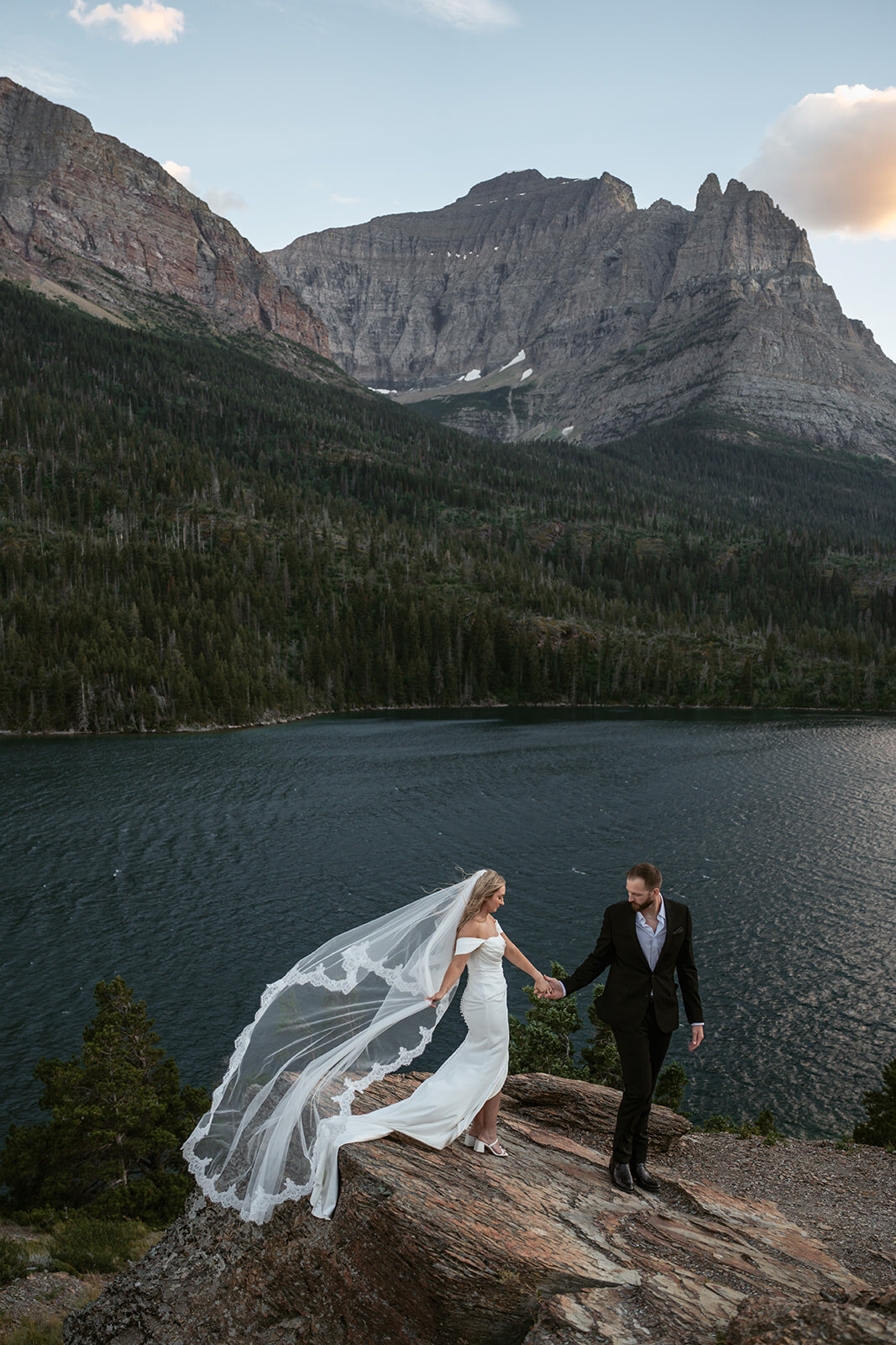A bride’s veil blows in the wind as she walks hand in hand with her groom along a rocky lakeside overlook surrounded by mountains in Glacier National Park, captured by Sydney Breann Photography.