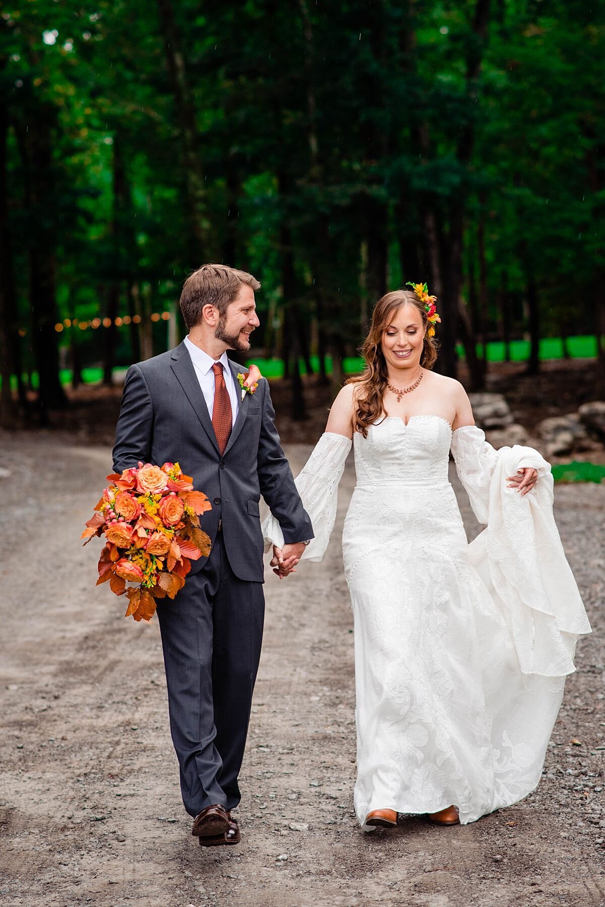 The groom, dressed in a charcoal gray suit, leads the bride, who is dressed in a detachable sleeve boho wedding dress, by the hand while carrying her bouquet of autumn leaves.