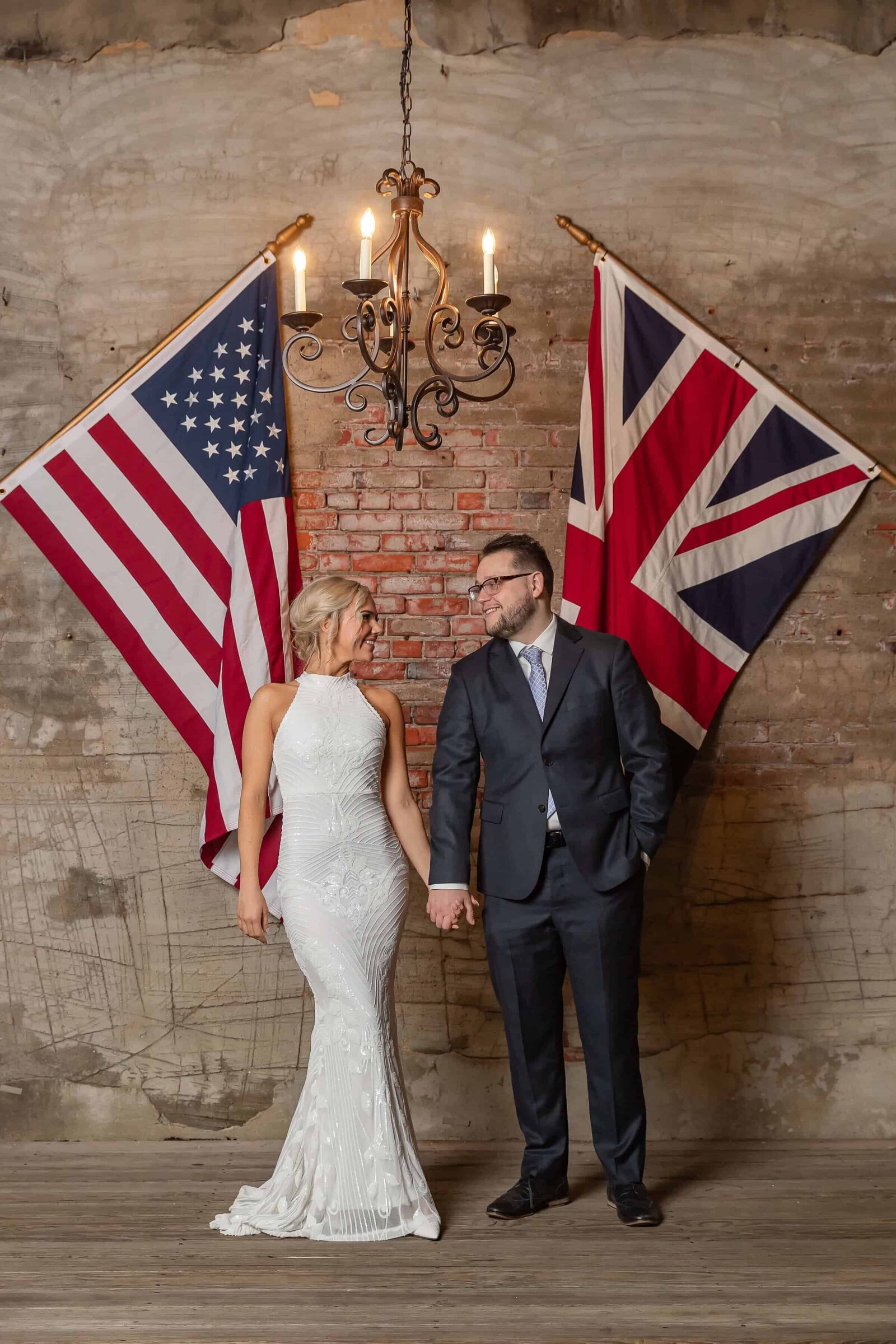A bride in a white dress and a groom in a dark suit hold hands and smile at each other, standing beneath a chandelier between a U.S. flag and a U.K. flag against a rustic brick wall.