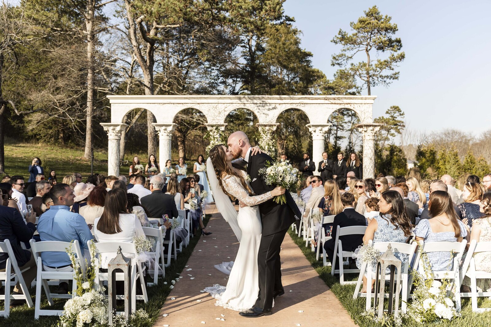 The bride and groom share a celebratory kiss at the end of the aisle, surrounded by cheering guests. The sun casts a golden glow over the elegant Greco-Roman-style columns framing their ceremony backdrop. The bride’s fitted lace wedding dress flows behind her as she clutches a bouquet of white florals, while the groom leans in, dressed in a sharp black tux